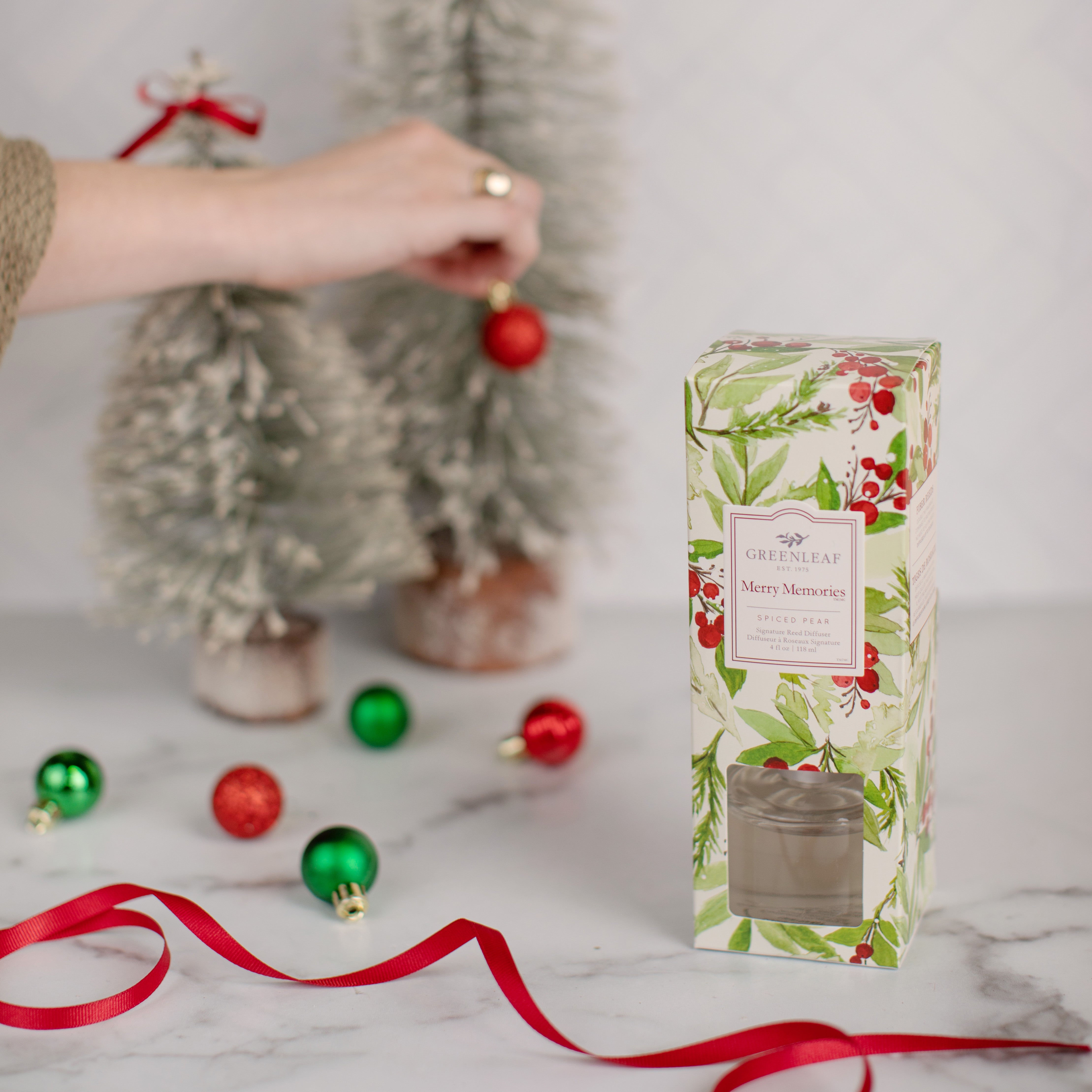 A hand hangs a red ornament on a frosted Christmas tree. Nearby, ornaments, a red ribbon, and a festive box labeled Greenleaf Gifts Merry Memories Reed Diffuser evoke holiday fragrance and cheer on the marble surface.