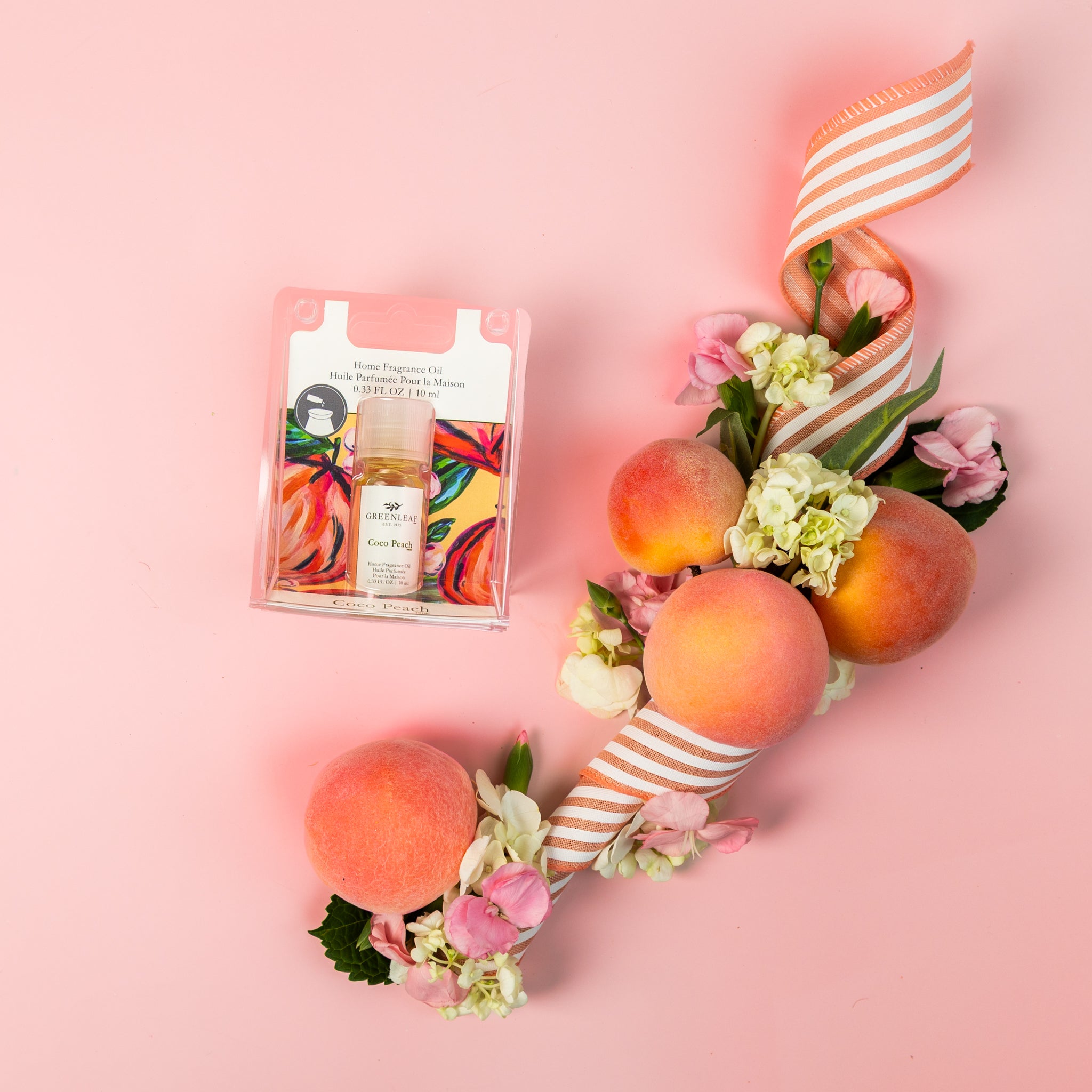 A flat lay on a pink background showcases Greenleaf Gifts' Coco Peach Home Fragrance Oil, fresh peaches, a striped ribbon, and assorted flowers arranged artfully.