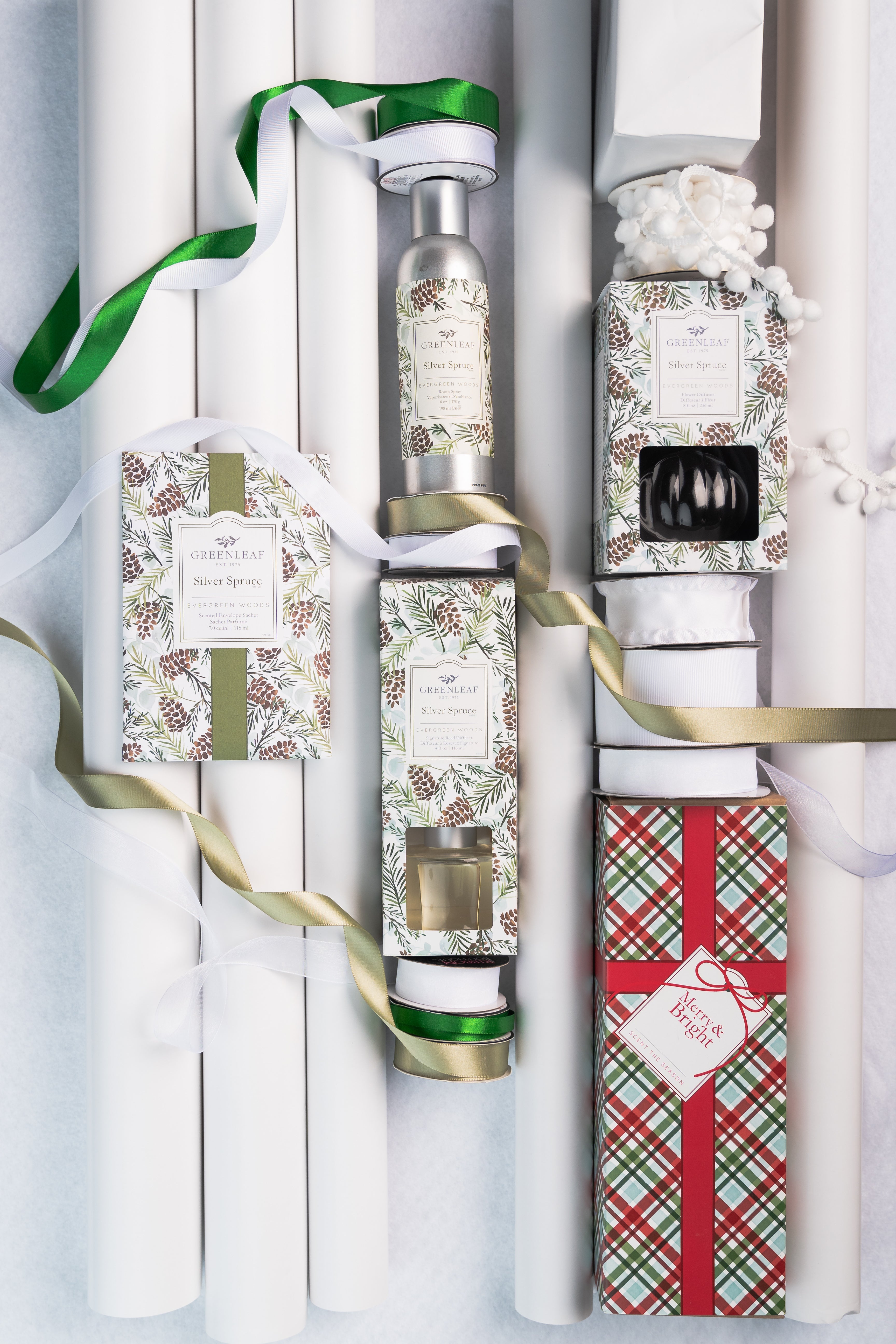 Gift items and wrapping supplies, including a Greenleaf Gifts Silver Spruce Reed Diffuser with fiber reeds, white paper rolls, pine/pinecone boxes, plaid gift box, small bottle, white ribbon, plus green and gold ribbons on a white surface.