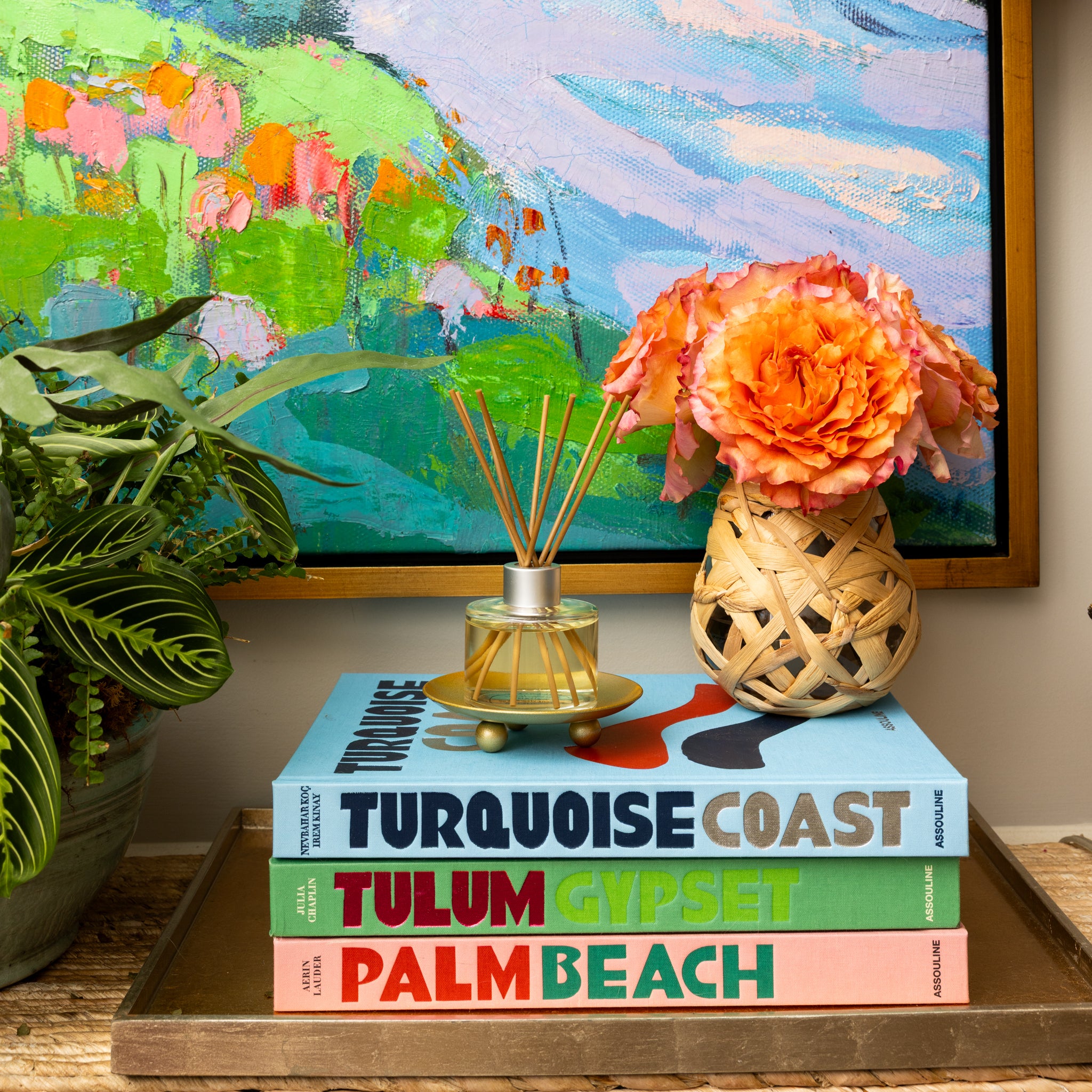 A small table holds colorful books—TURQUOISE COAST, TULUM GYPSET, PALM BEACH—plus a Greenleaf Gifts Coco Peach Reed Diffuser, an orange flower arrangement in a woven vase, a potted plant, and a bright painting.