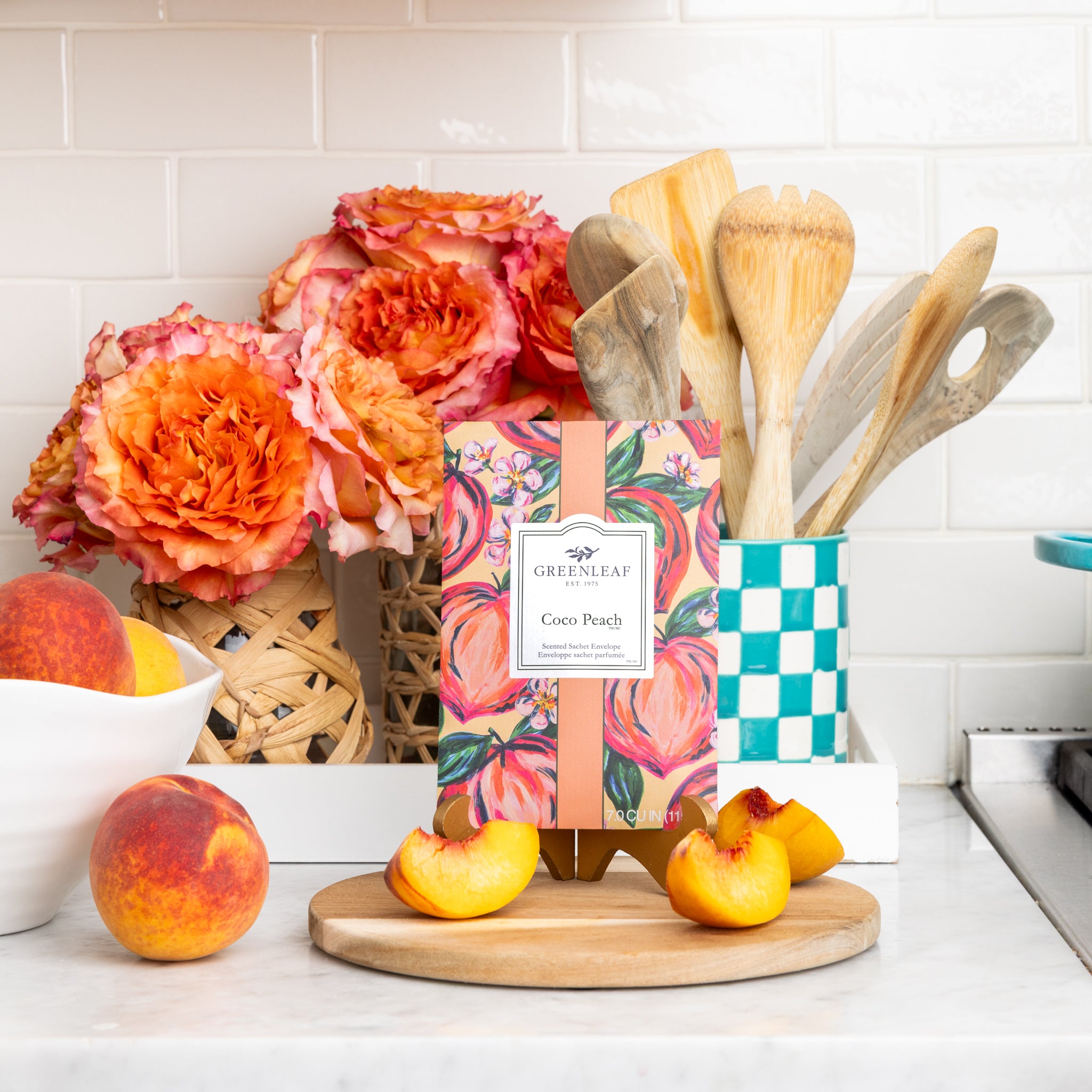 A box of Greenleaf Gifts Coco Peach Sachets sits on a wooden board with sliced peaches, fresh peaches, bright orange flowers, and a container of wooden utensils, filling the air with a long-lasting cocoa peach scent.