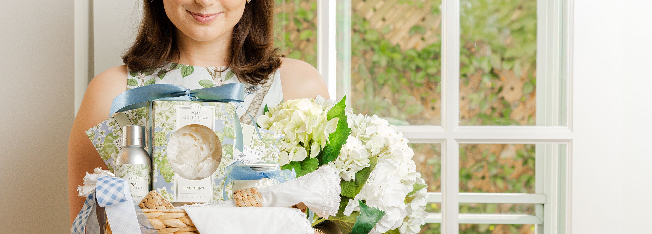 Woman holding a gift basket filled with Greenleaf Hydgrangea products and flowers in a bright room.