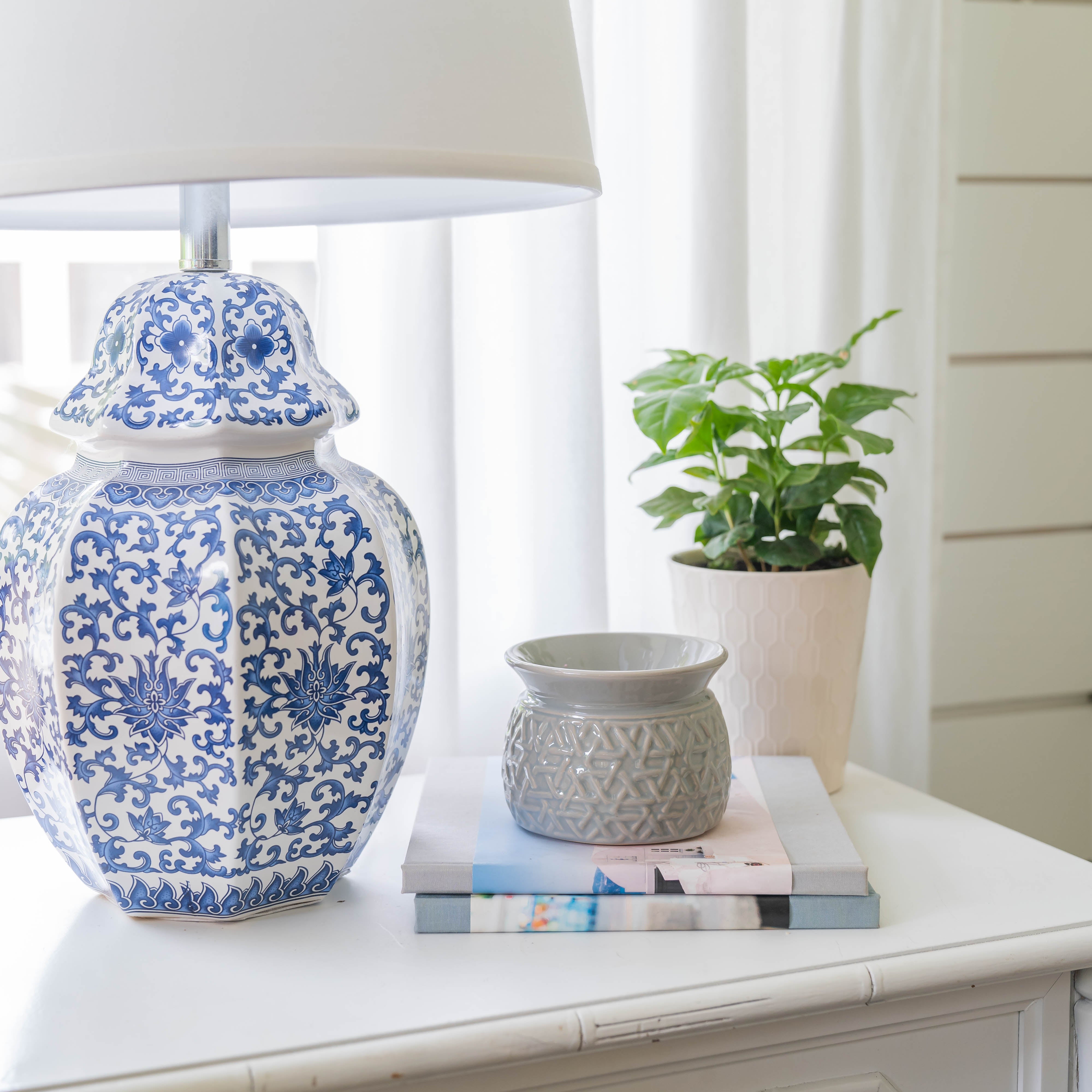A white nightstand with a blue and white patterned lamp, small gray vase, stacked books, and a potted green plant is complemented by the gentle rose scent of Greenleaf Gifts’ Roses Home Fragrance Oil—set against white curtains and walls.