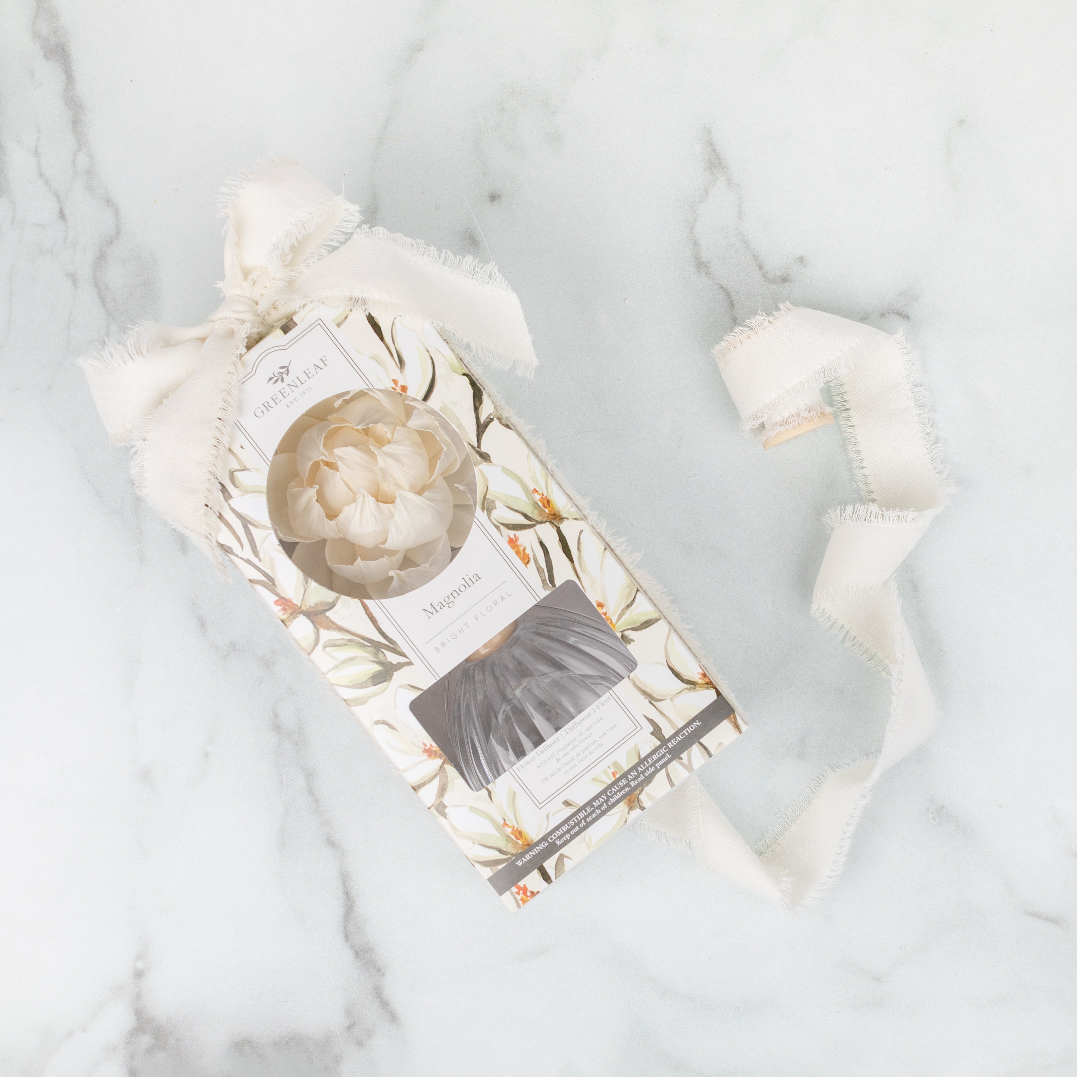 A Greenleaf Gifts Magnolia Flower Diffuser box, adorned with a white peony illustration and label, rests on marble. Wrapped in a frayed white ribbon, it hints at the soothing fragrance oil and diffuser inside.