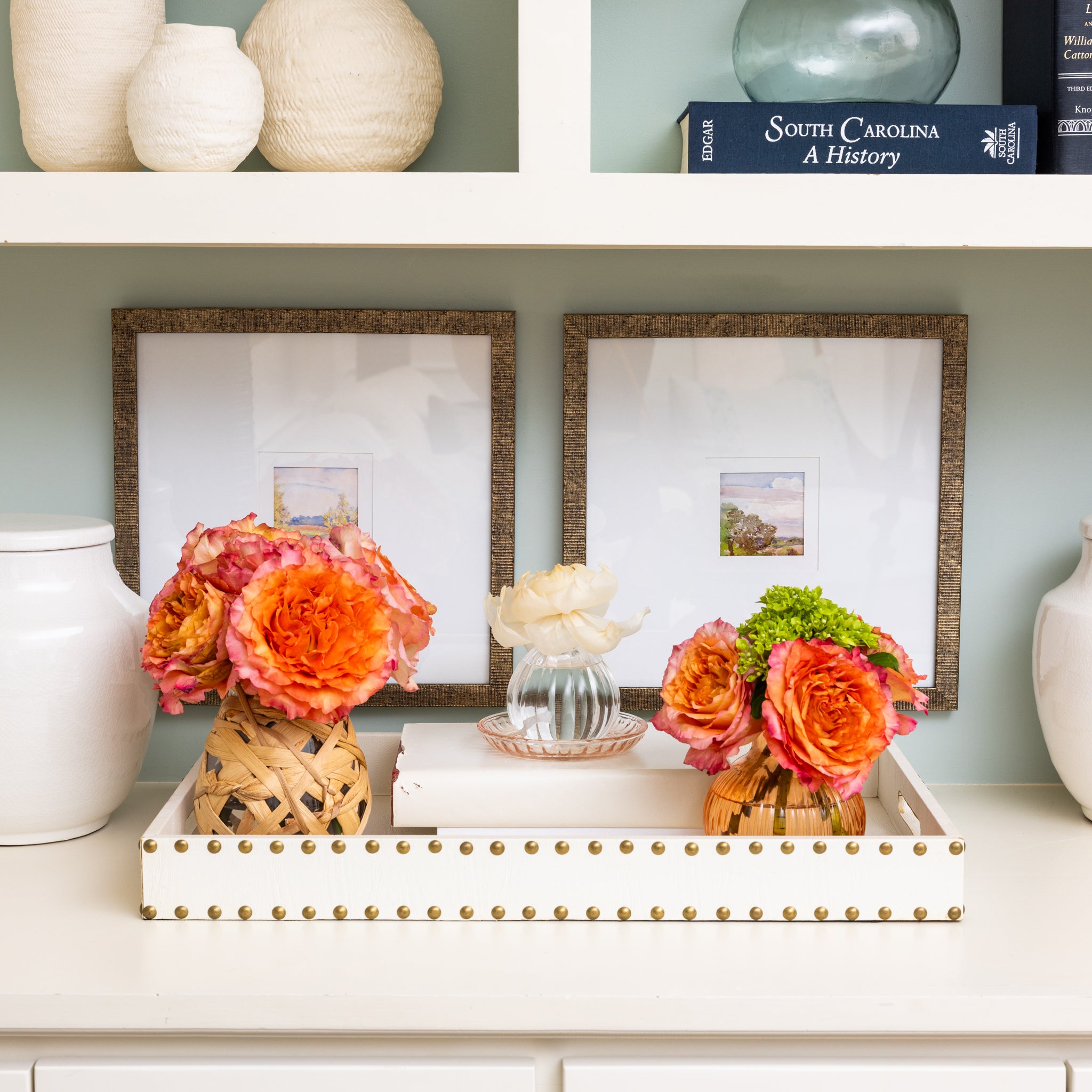 A white shelf displays the Greenleaf Gifts Coco Peach Flower Diffuser, orange and white flowers with a sola paper flower, framed art, white ceramic vases, and books—including “South Carolina: A History.”.