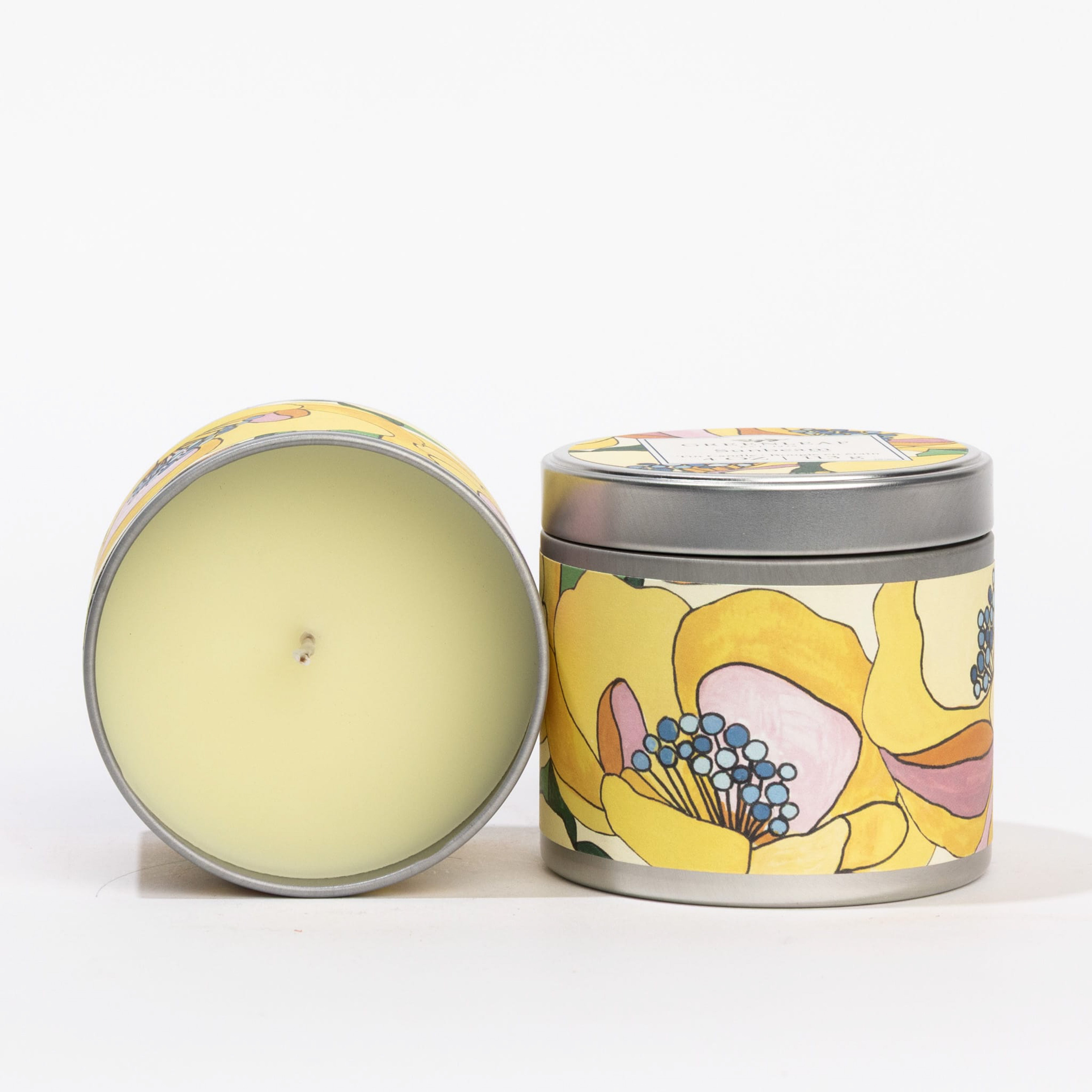Two Sunbeam Tin Candles by Greenleaf Gifts—one upright with a vibrant hand-painted lid, the other on its side, showing the candle wick and colorful floral tin design.