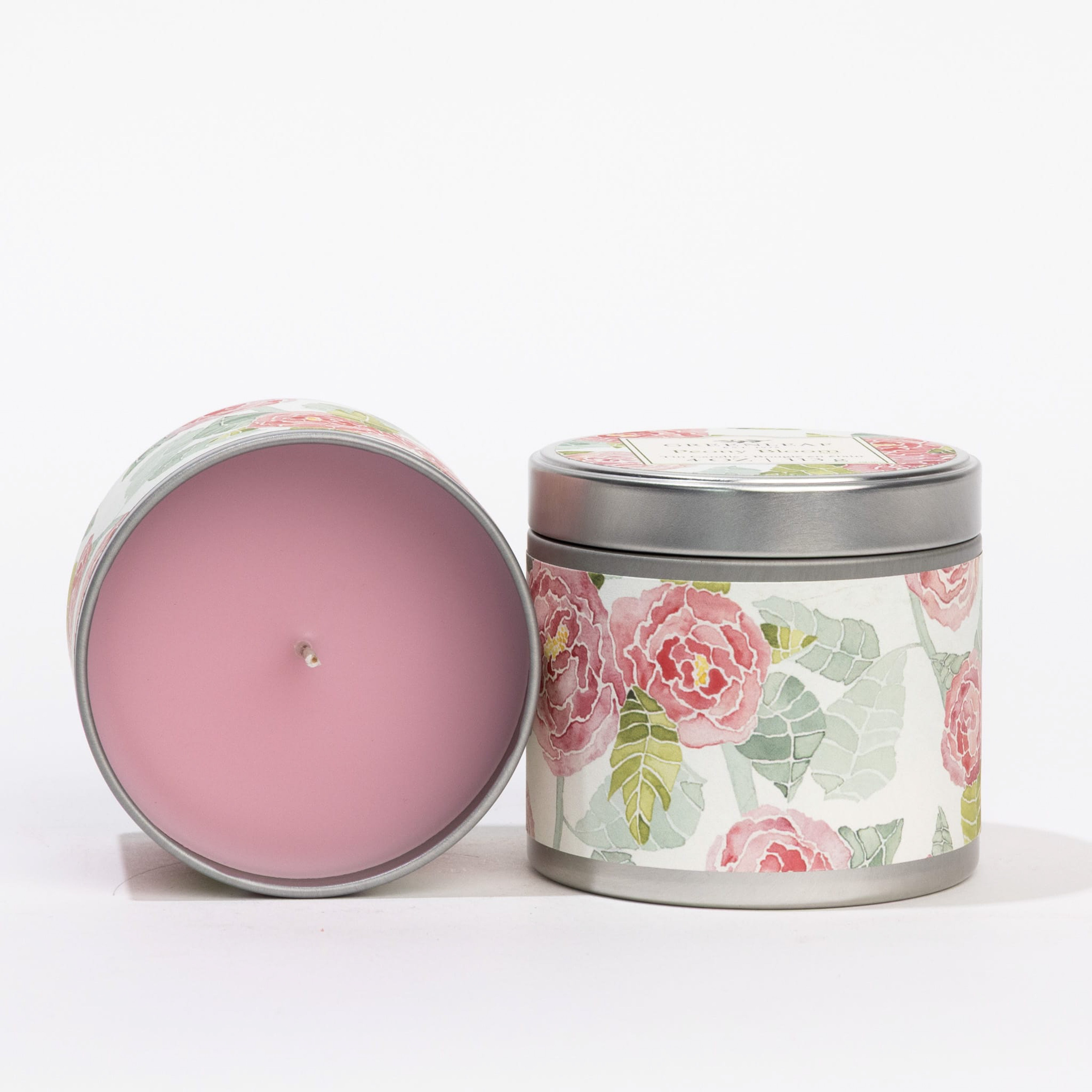 Two Greenleaf Gifts Peony Bloom Tin Candles are shown: one closed and upright, the other open to display a pink candle. Both tins feature pink flower and green leaf designs on white backgrounds.
