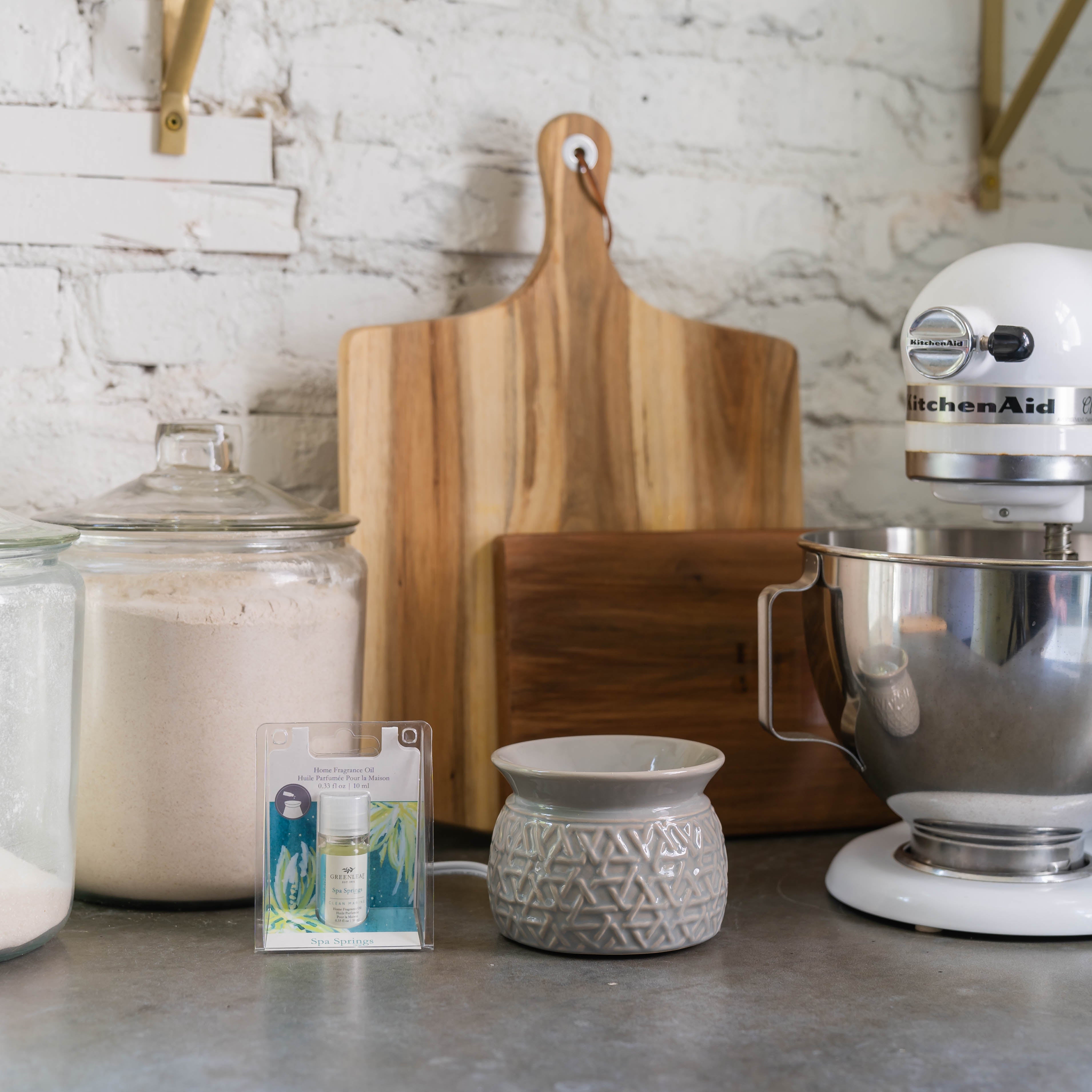A kitchen counter displays a stand mixer, a wood cutting board, jars of flour and sugar, a ceramic fragrance warmer, and Greenleaf Gifts Spa Springs Home Fragrance Oil—all set against a white brick wall.