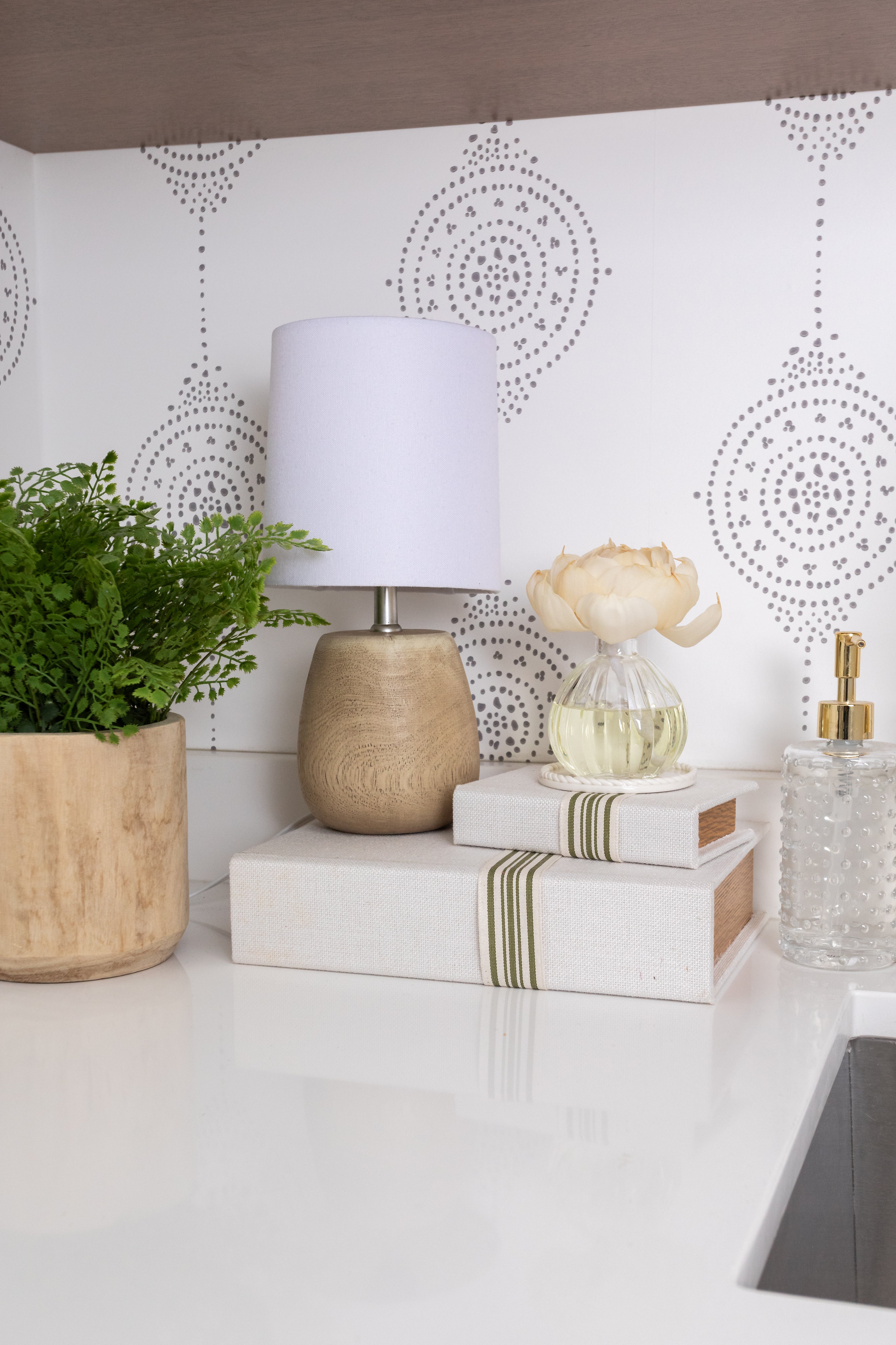 A small lamp with a white shade, a potted green plant, two stacked books, and the Willow & Sage Flower Diffuser by Greenleaf Gifts sit on a white countertop against a patterned white tile backsplash.