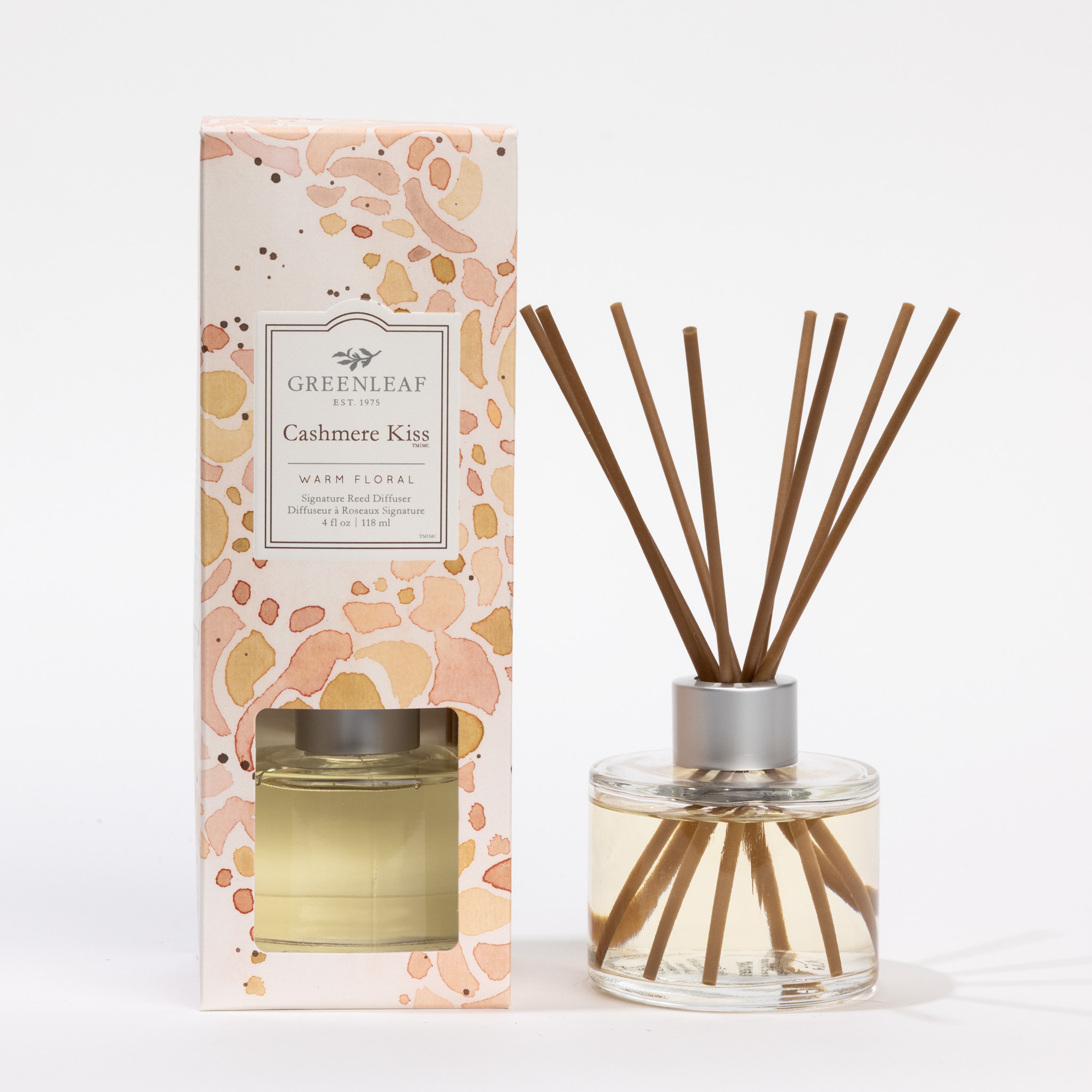 The Greenleaf Gifts Cashmere Kiss Reed Diffuser features fiber reeds in a clear glass bottle and comes in a patterned box with warm orange and pink floral accents, adding a touch of elegance to any space.
