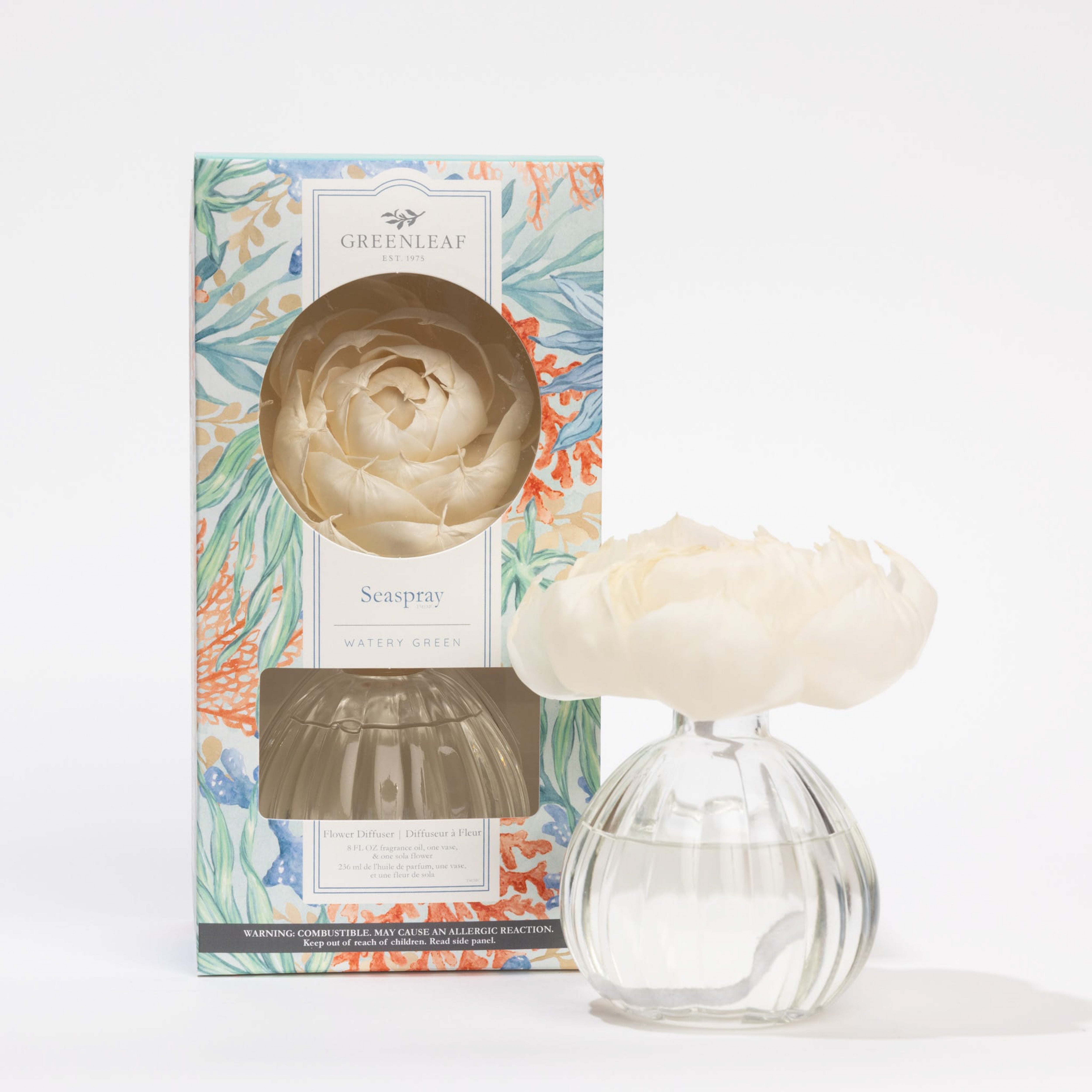 A clear glass vase with water holds a white artificial flower. Next to it is a Greenleaf Gifts Flower Diffuser-Seaspray box with floral patterns, a window displaying the diffuser, and fragrance lasting up to 60 days.