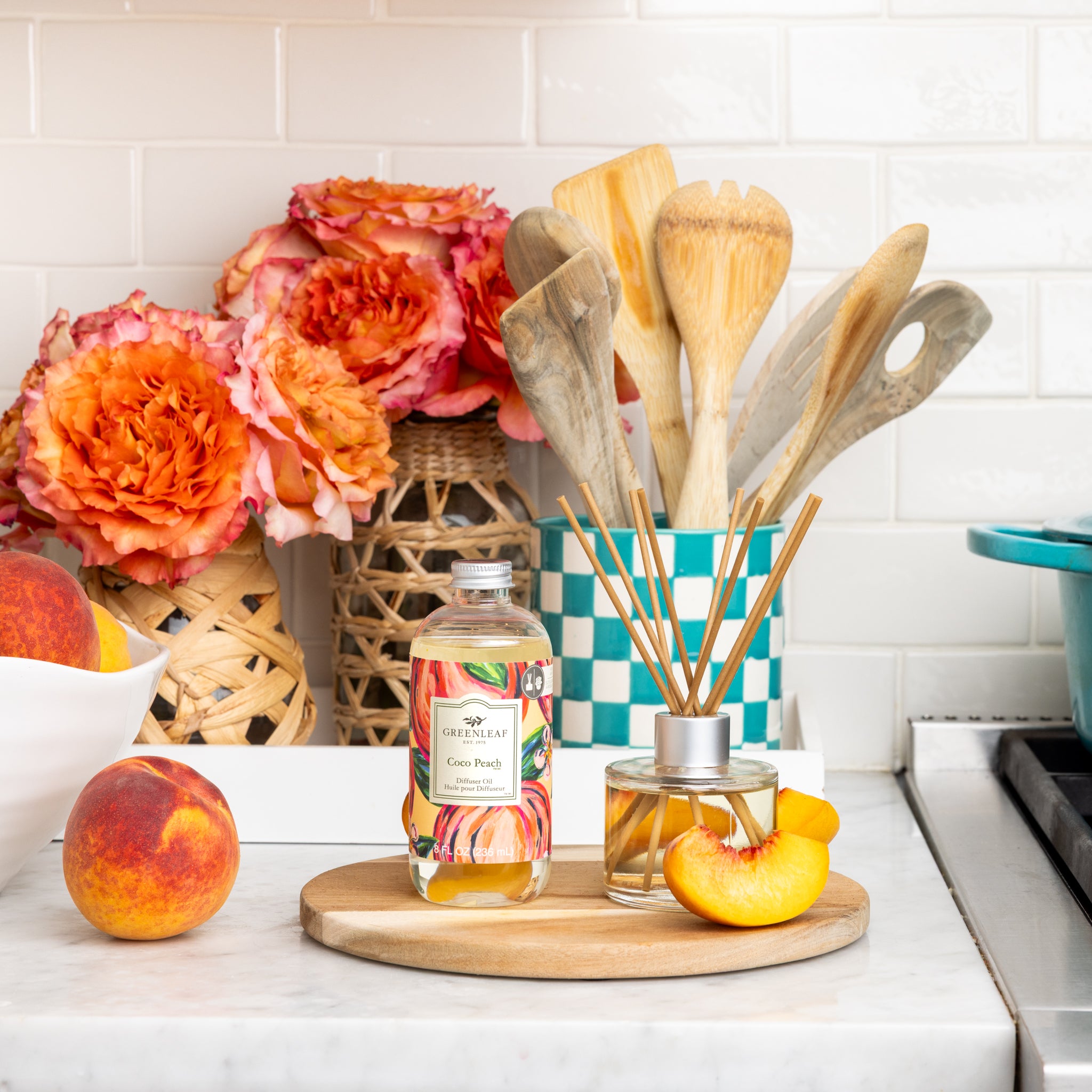 A kitchen counter with a bottle of Coco Peach Diffuser Oil by Greenleaf Gifts, a Greenleaf Flower Diffuser, fresh peaches and slices on a wooden tray, a checkered utensil holder with wooden spoons, and a vase of vibrant orange flowers.