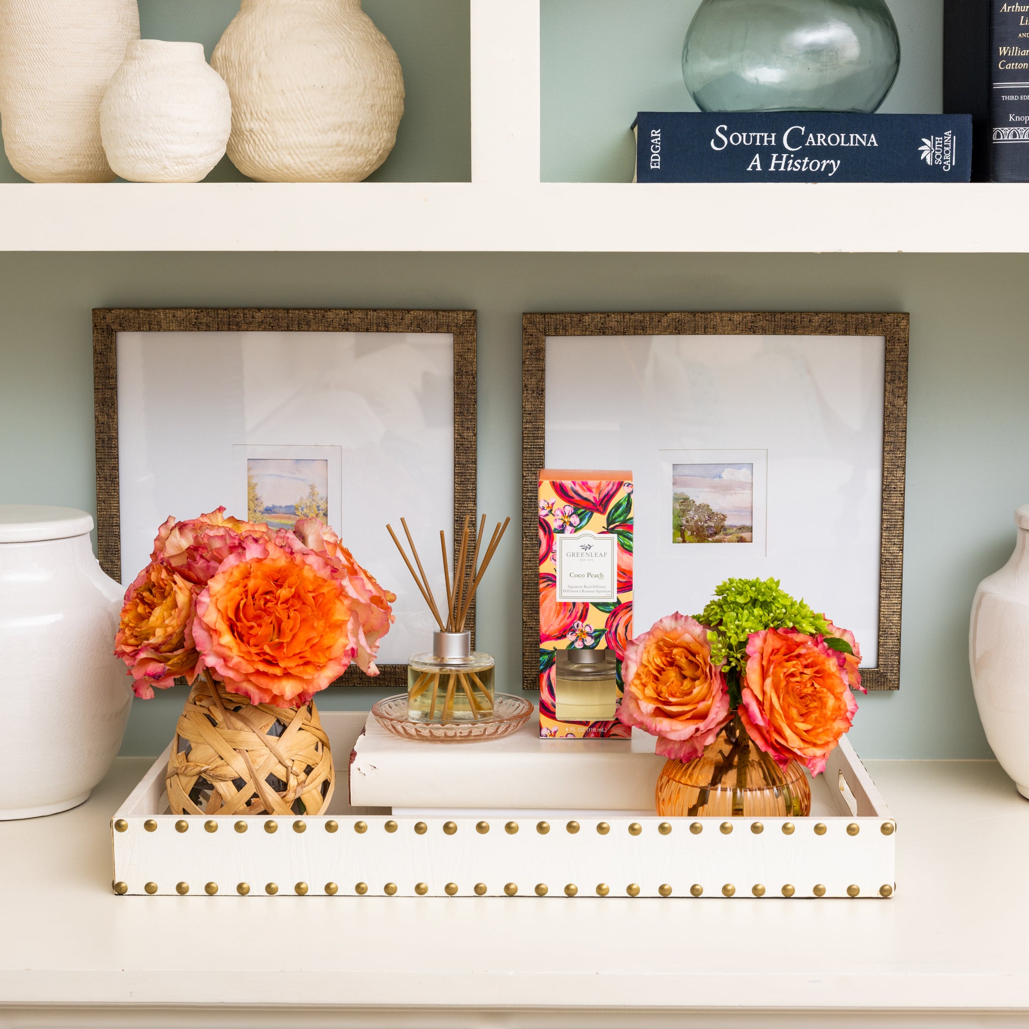 A white shelf displays vases, framed art, a blue book, and a tray with orange flower arrangements, the Greenleaf Gifts Coco Peach Reed Diffuser with fiber reeds, and a colorful box in a bright, stylish setting.