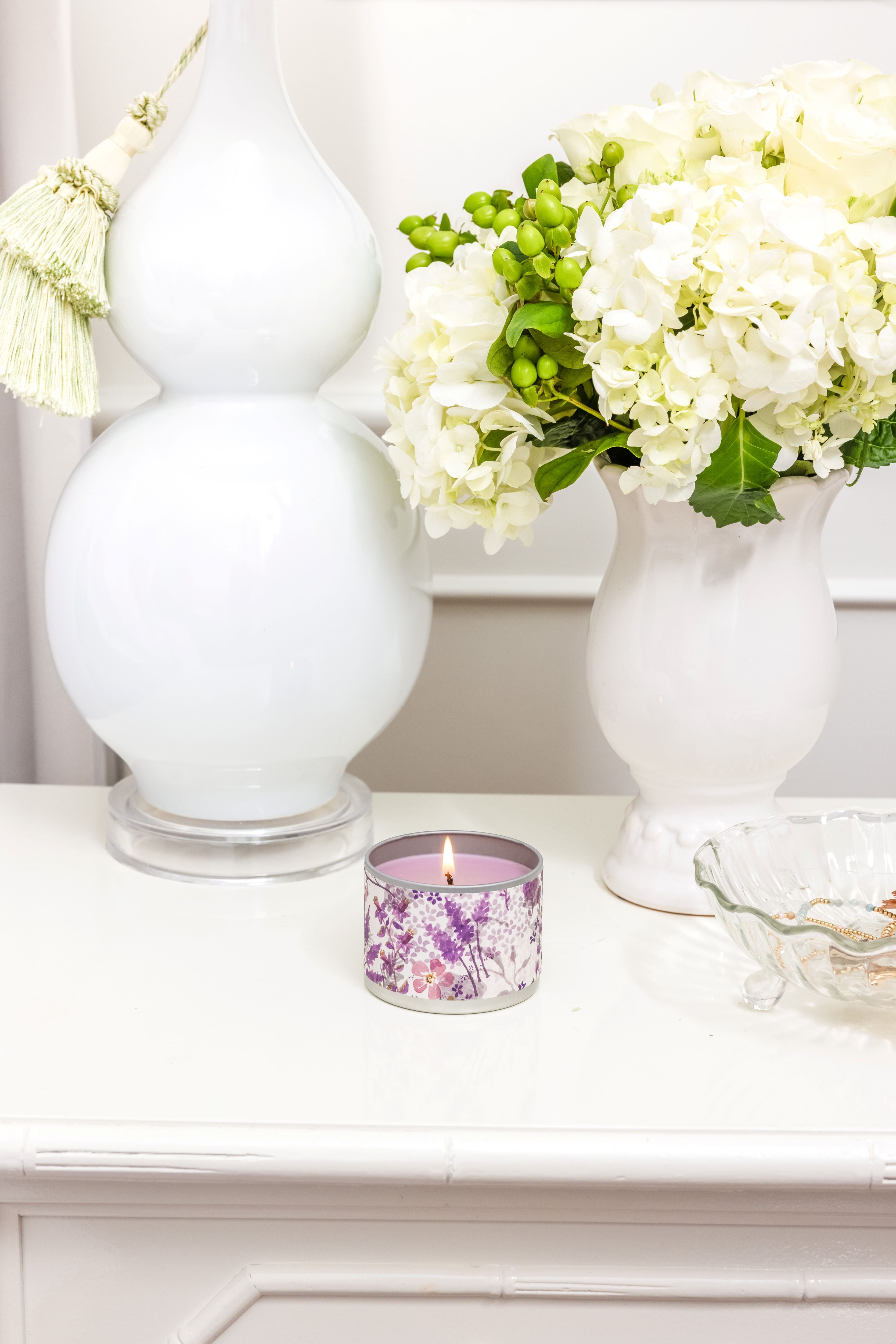 A lit Greenleaf Gifts Lavender Tin Candle with a floral scent sits on a white table near a vase of hydrangeas, a glass lamp with a tassel, and a decorative glass dish.