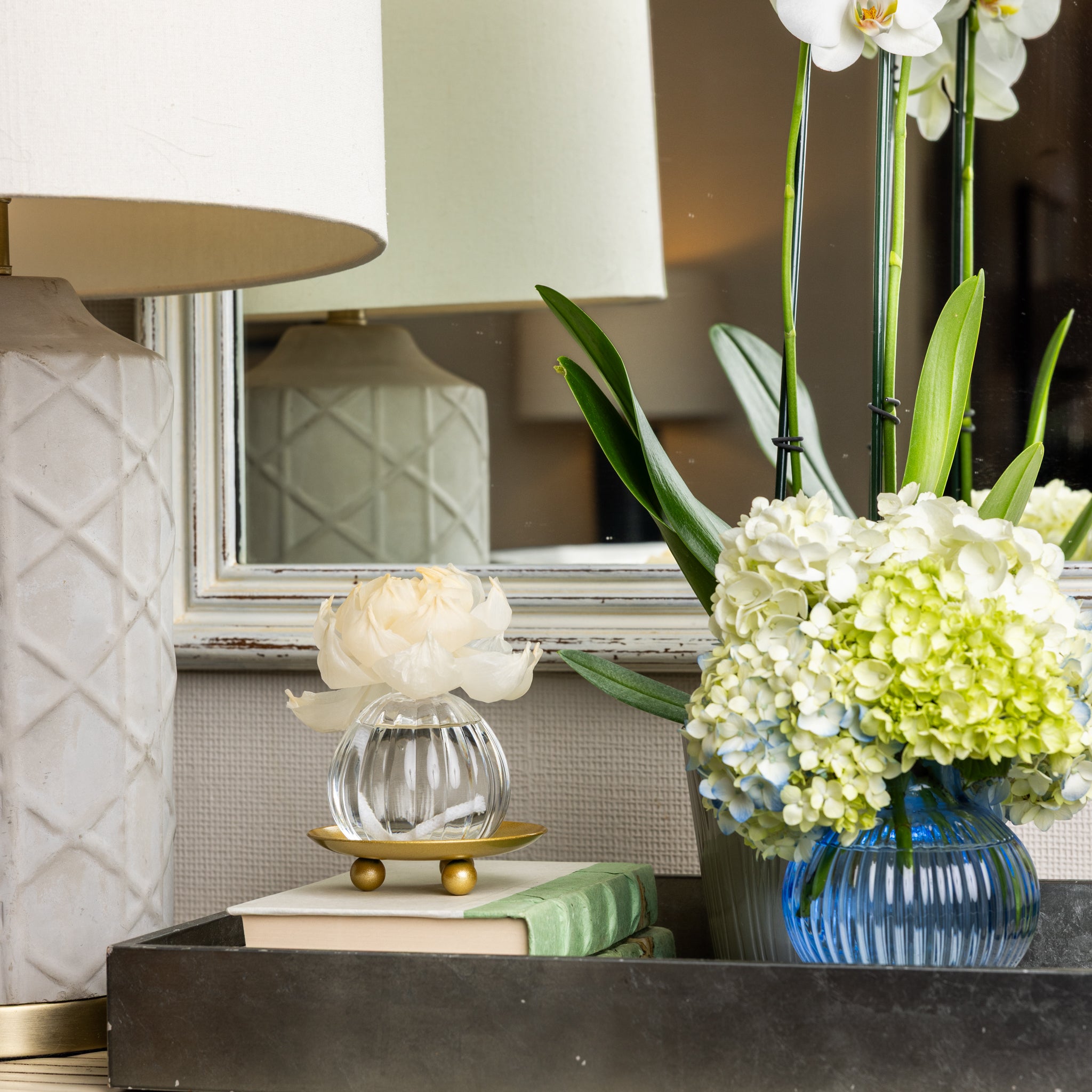 A tray on a table holds stacked books, a Greenleaf Gifts Waterplants Flower Diffuser with a sola paper flower, and a blue vase with white hydrangeas and orchids. A lamp and mirror are in the background.