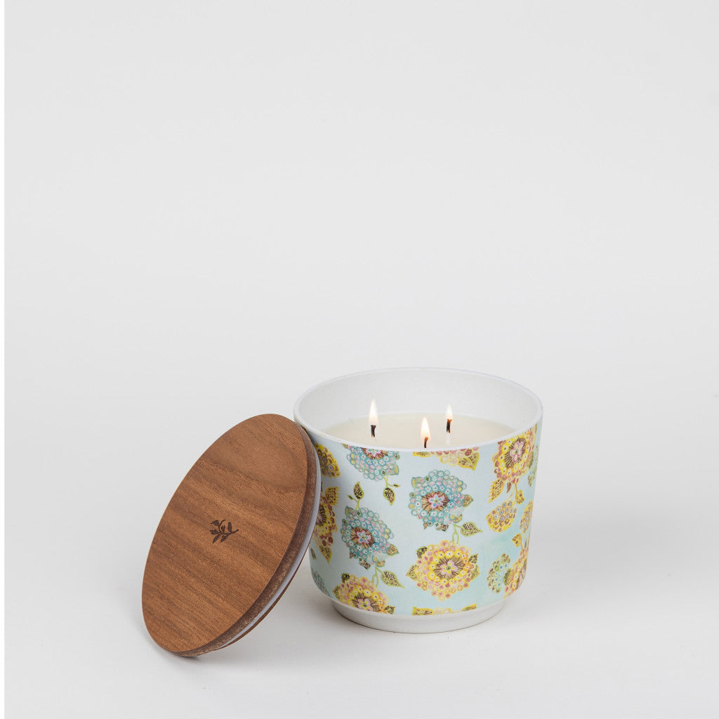 The Greenleaf Gifts Patterned 3 Wick Candle - Bella Freesia features a floral scent and glows in a light blue, floral-patterned container with a wooden lid resting beside it, all set against a plain white background.