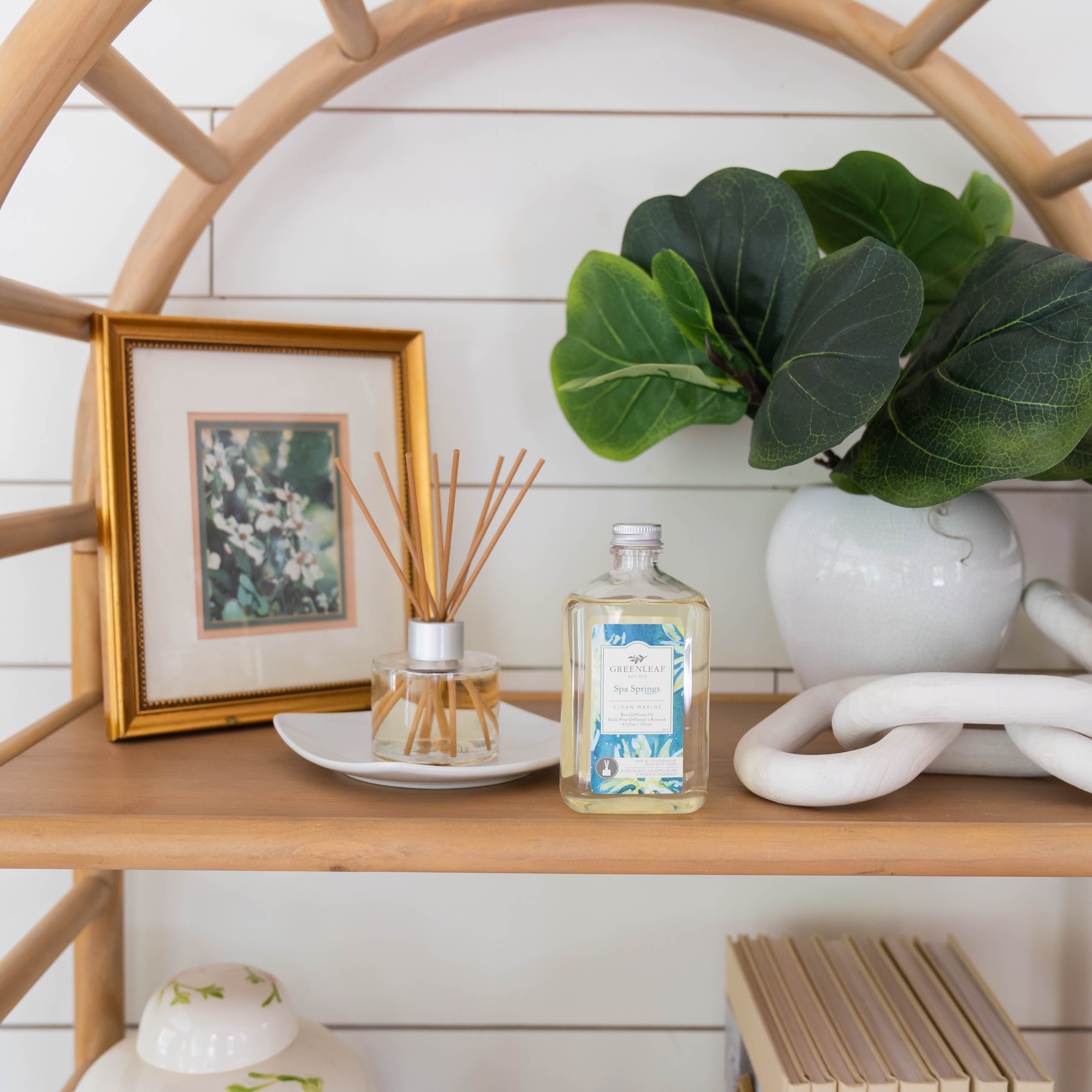 A wooden shelf displays a gold-framed floral photo, a bottle of room spray, a white vase with green leaves, a decorative ceramic chain, and Greenleaf Gifts’ Spa Springs Diffuser Oil Refill to refresh your space with calming fragrance.