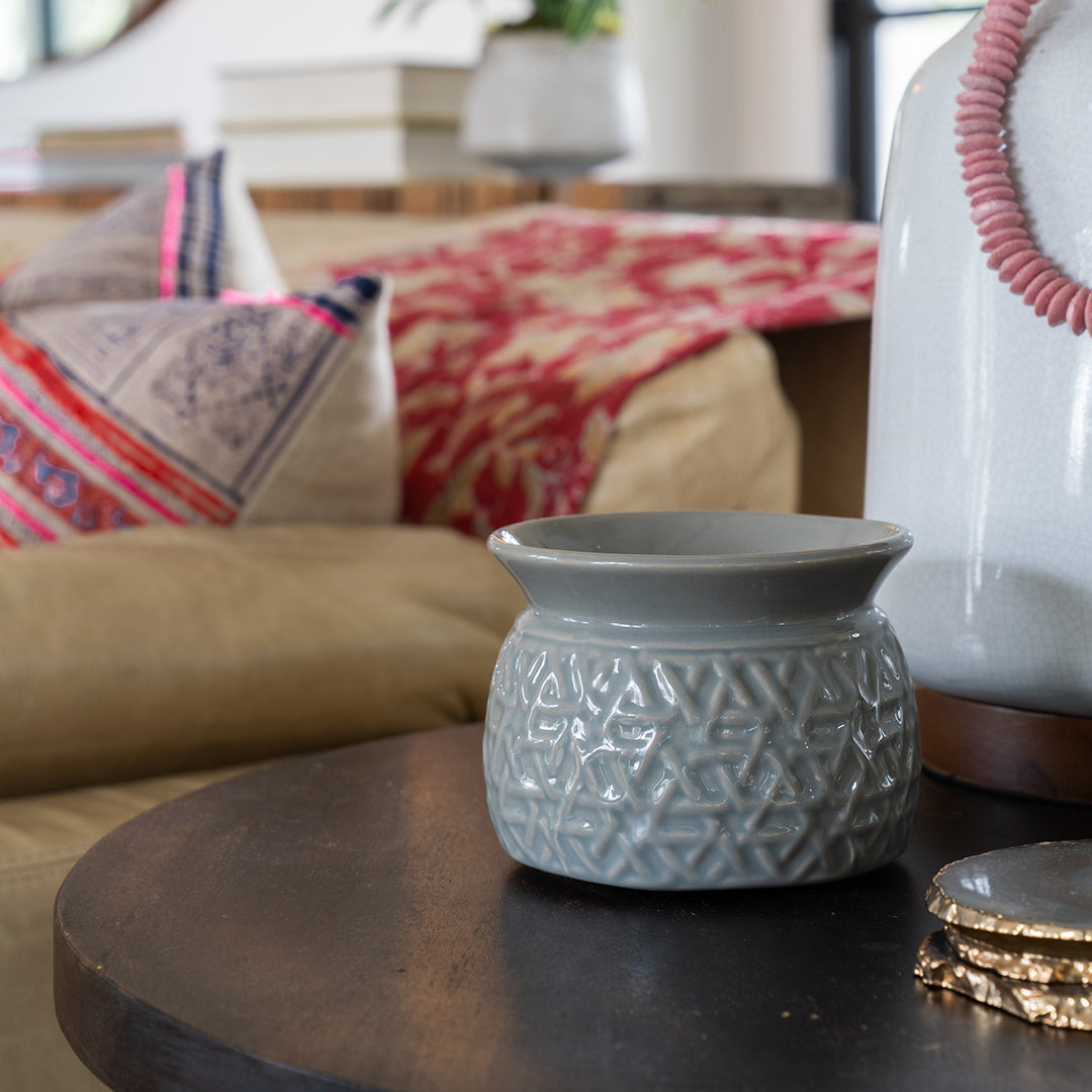 A gray ceramic pot with a geometric pattern sits on a dark wooden table, as the soothing scent of Greenleaf Gifts Roses Home Fragrance Oil fills the air. In the background, a beige couch with patterned pillows and stacked books adds warmth.