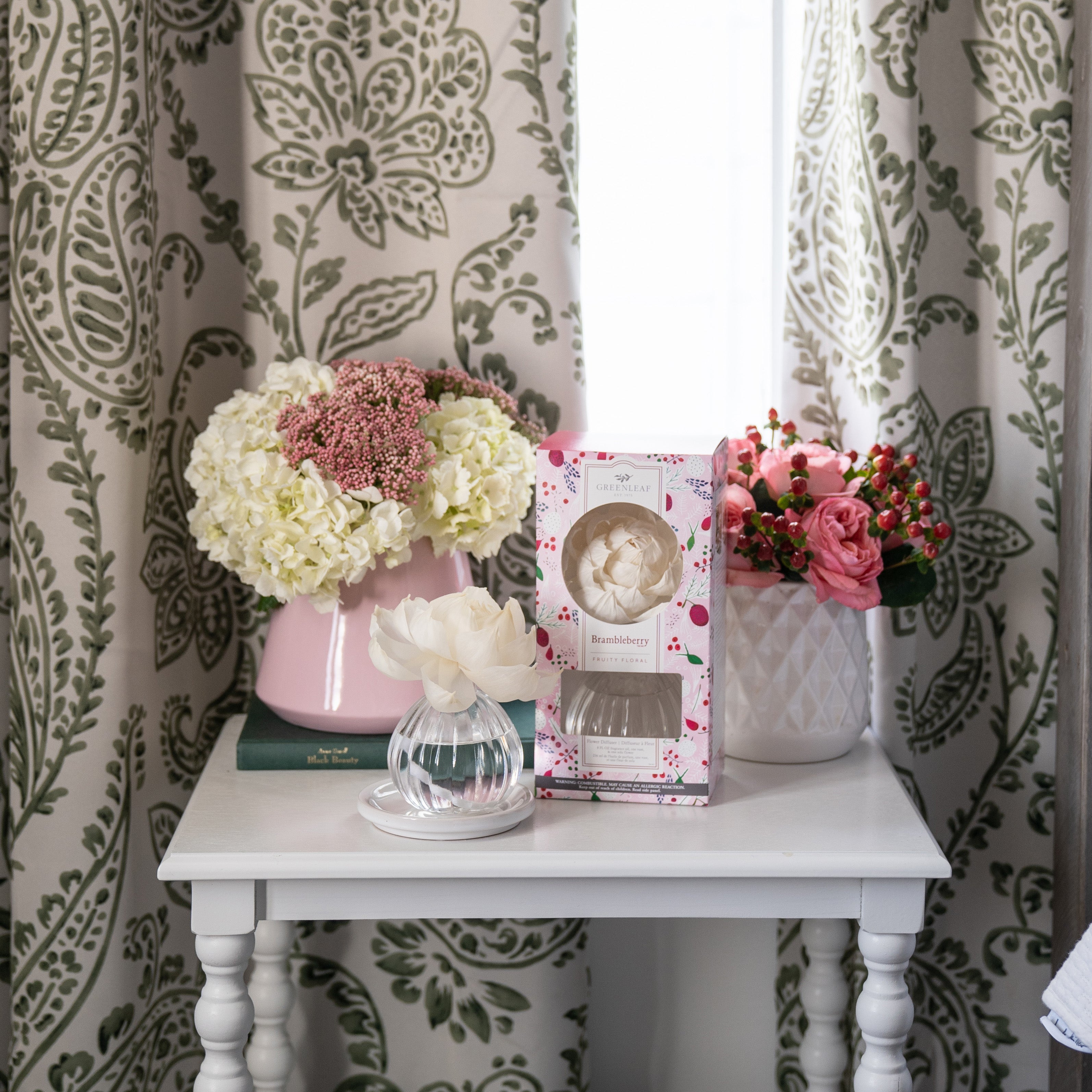 A white side table with books, flower arrangements in pink and white vases, a Greenleaf Gifts Brambleberry Flower Diffuser (8 oz), set against green and white paisley curtains. Style code GLG966572S. Returns within 60 days available.