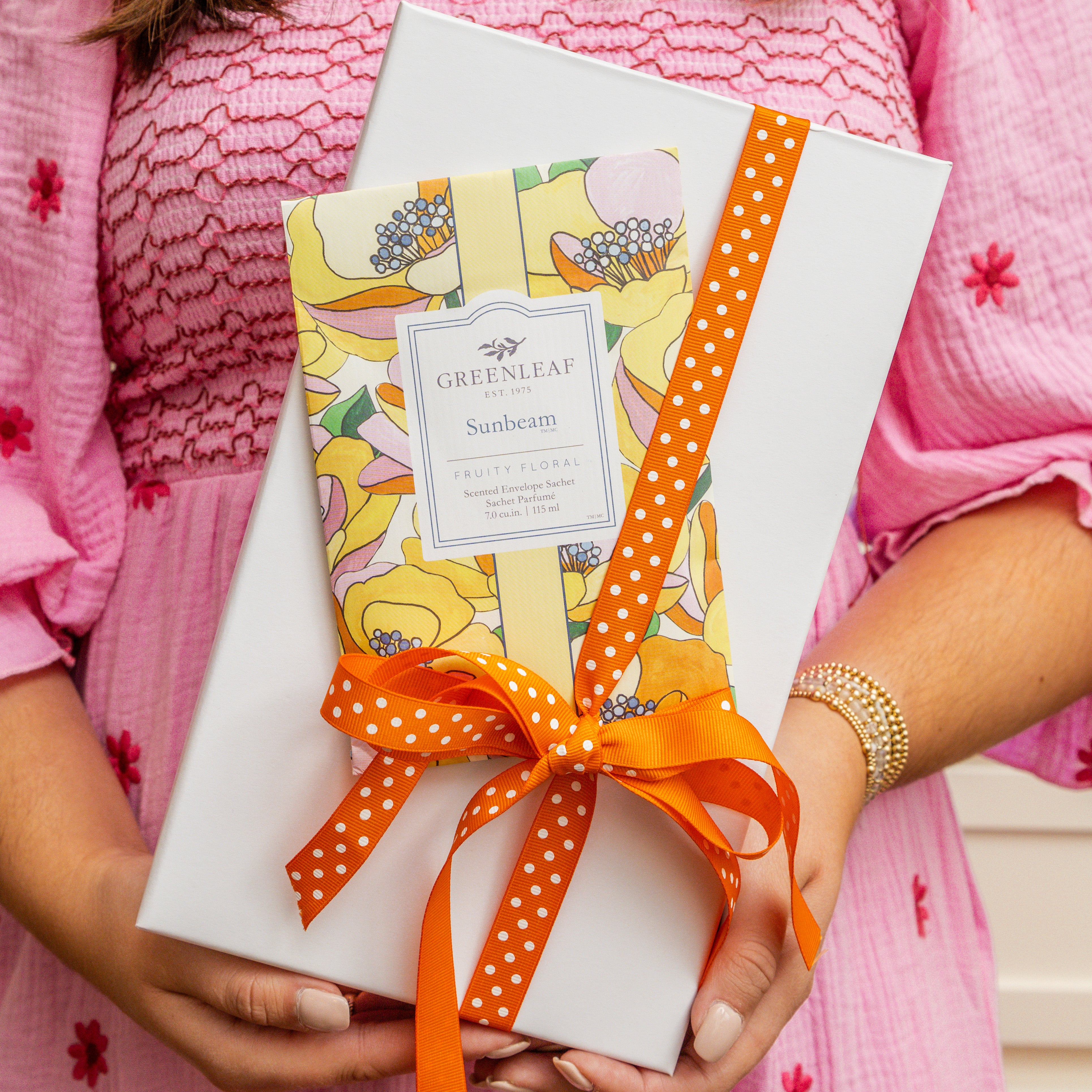 A person in a pink dress holds a white gift box with an orange polka dot ribbon and a Greenleaf Gifts Sunbeam Sachet, known for its long-lasting fragrance, attached to the top.