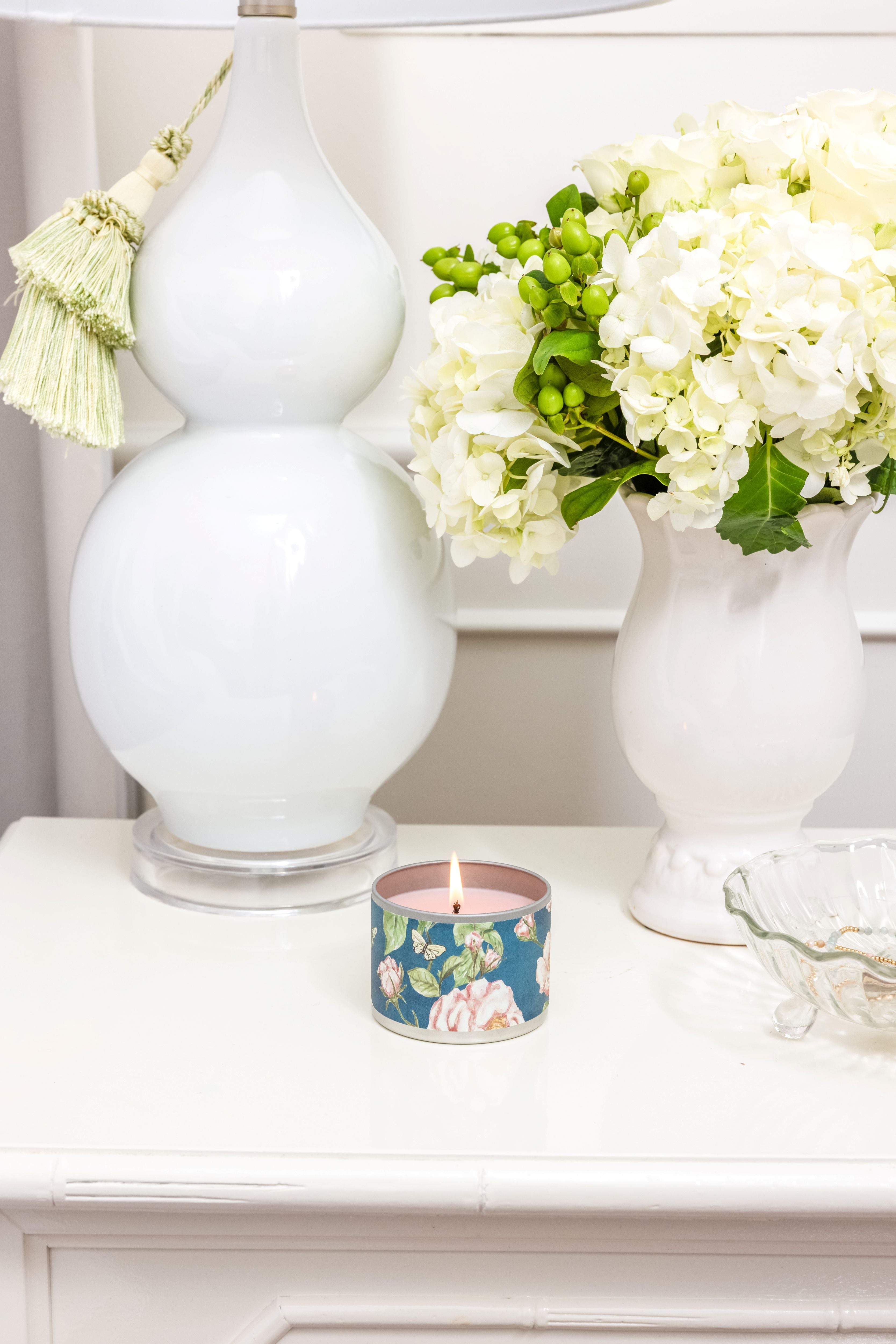 A Greenleaf Gifts Roses Tin Candle sits on a white nightstand next to a white table lamp, a vase of white hydrangeas with green buds, and a glass dish.