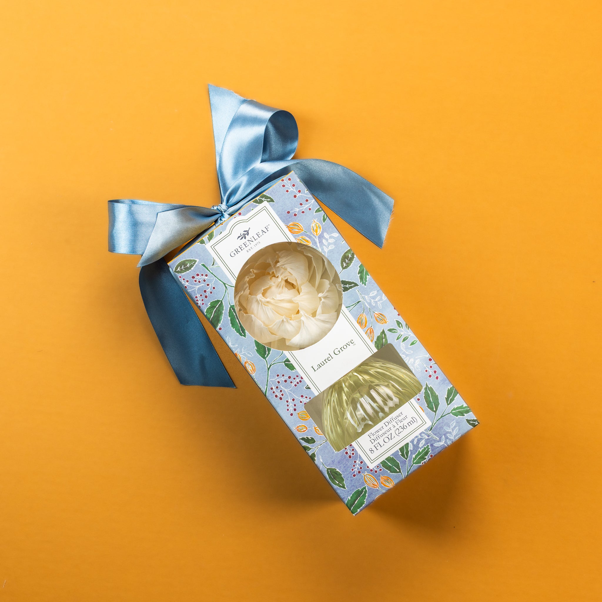 A blue floral-patterned gift box with a satin blue ribbon and a white flower-shaped diffuser is displayed on a yellow background. The label reads Greenleaf Gifts, Laurel Grove Flower Diffuser.