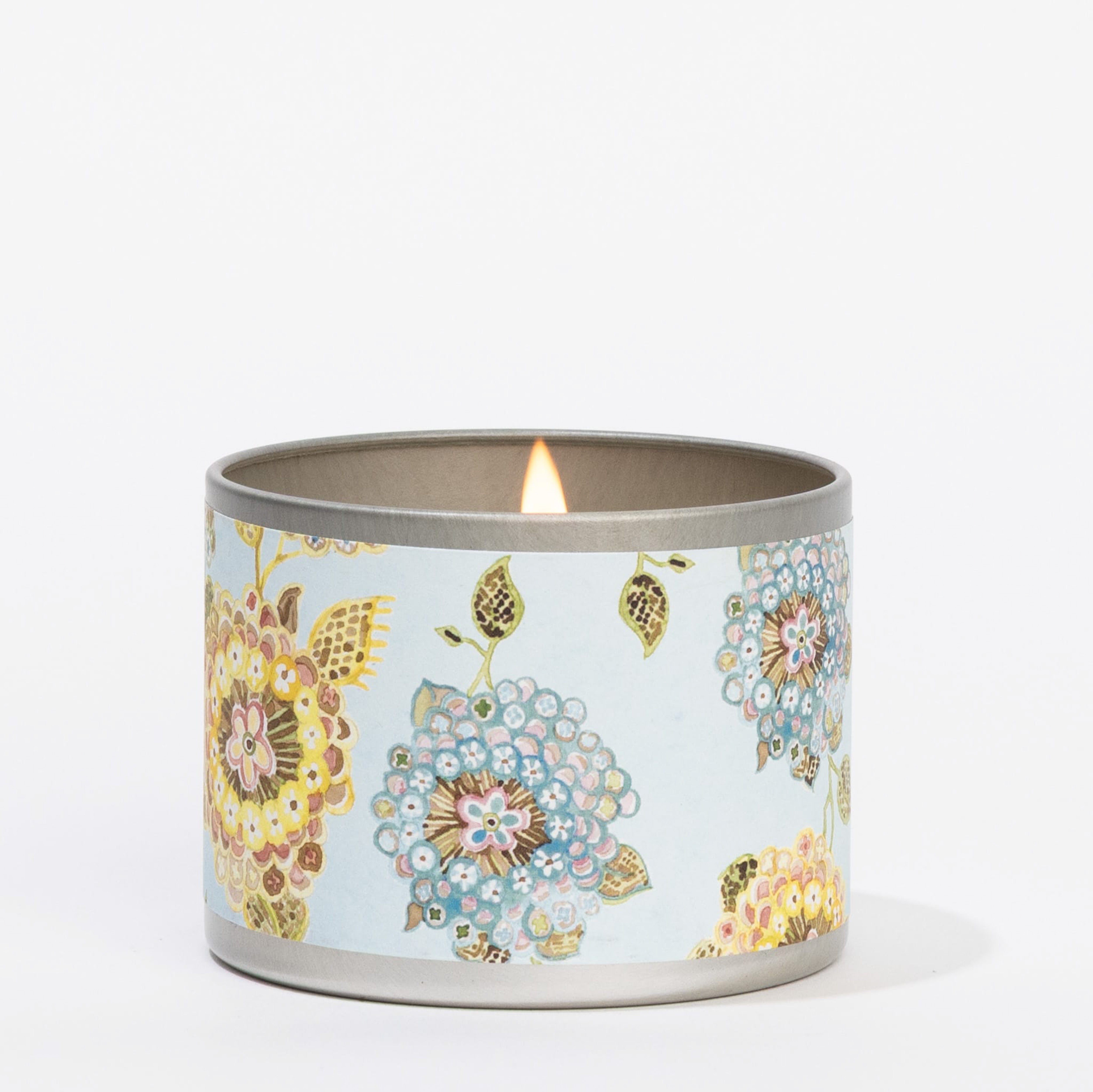 The Bella Freesia Tin Candle by Greenleaf Gifts sits on a white surface; its round metal container features a light blue background with colorful floral patterns and offers a refreshing Bella Freesia scent.