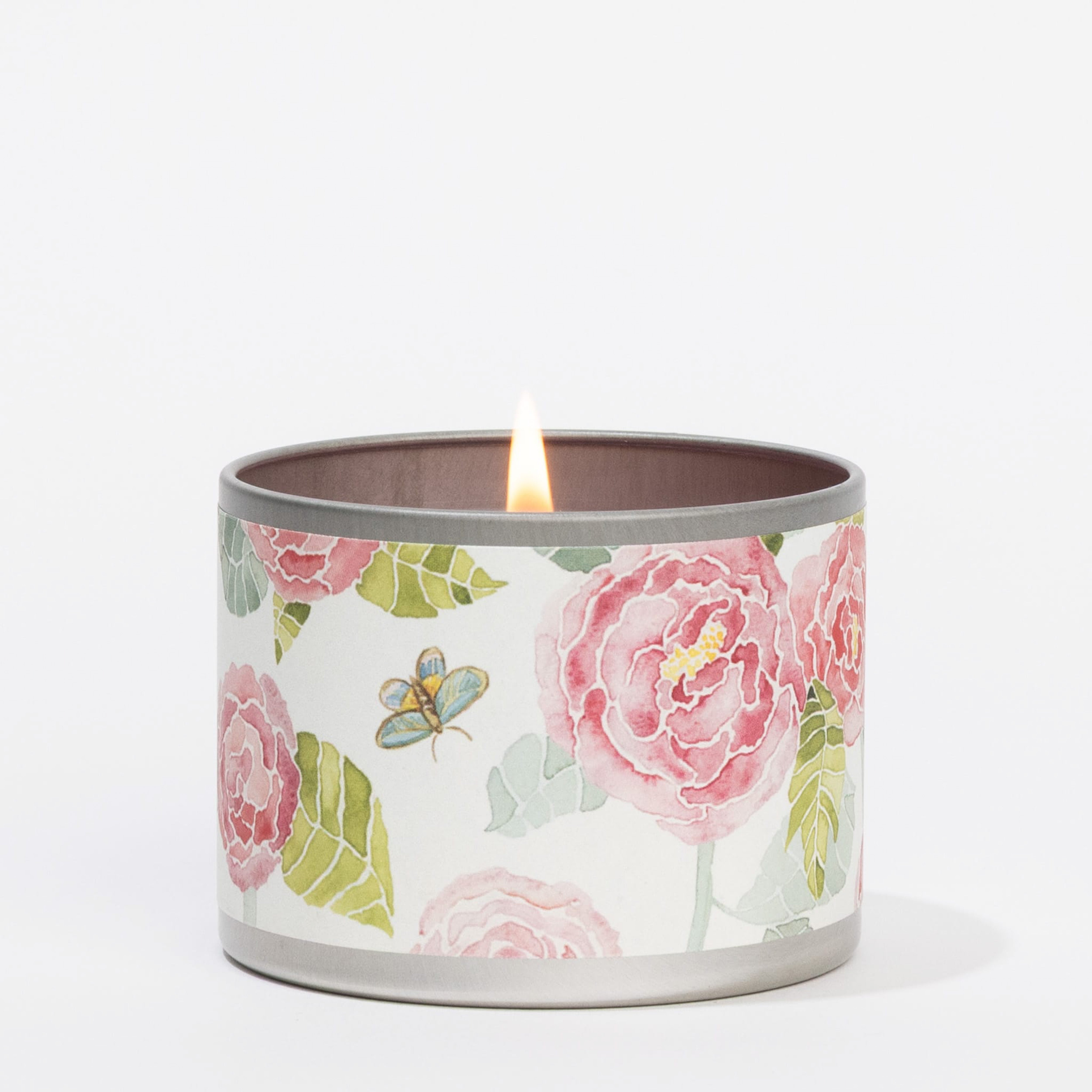 The Peony Bloom Tin Candle by Greenleaf Gifts, in a round tin adorned with pink flowers, green leaves, and a butterfly design, sits elegantly on a white surface—perfect as a portable candle for any occasion.