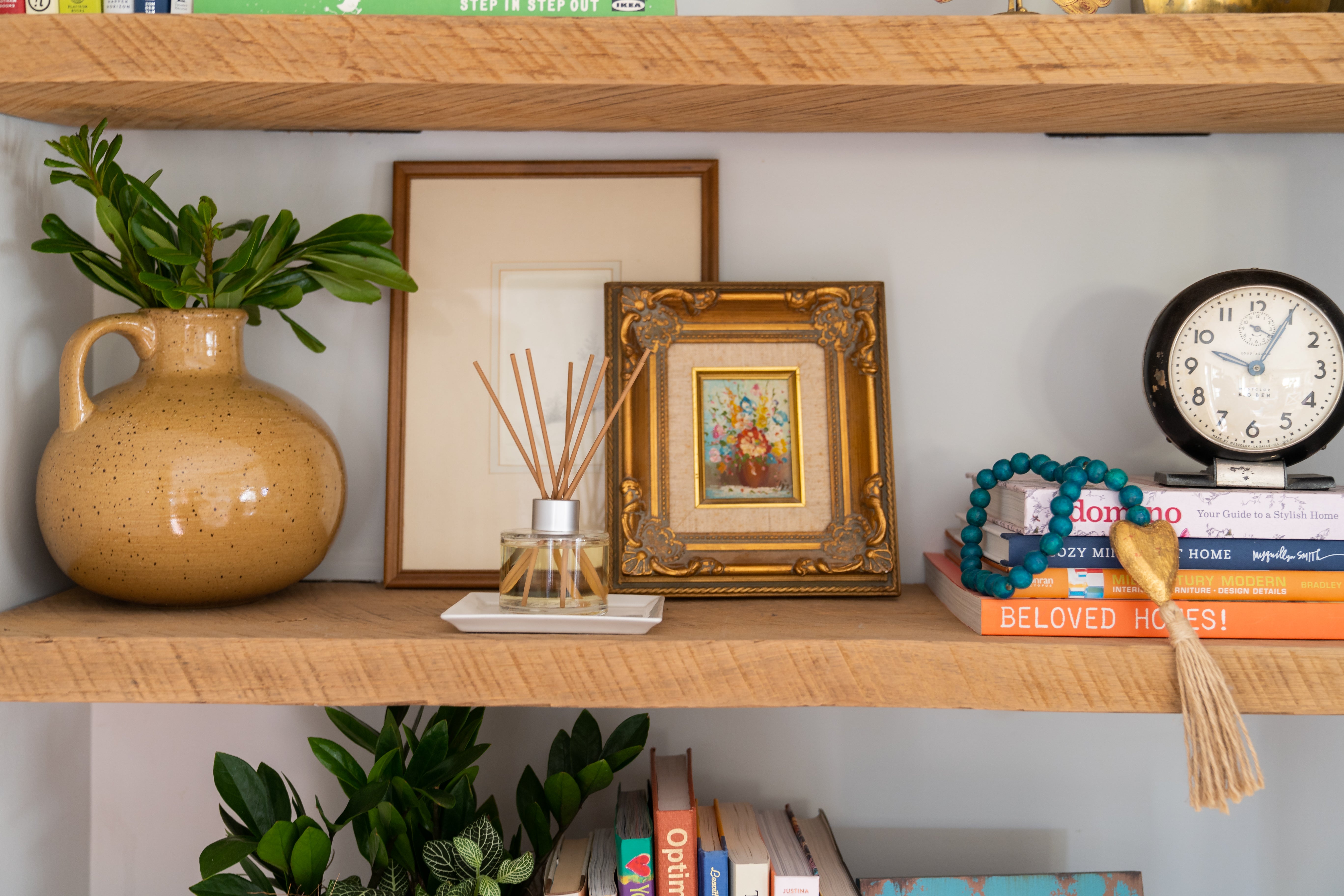 A wooden shelf showcases a beige vase with green leaves, a Greenleaf Gifts Spa Springs Reed Diffuser with fiber reeds, two framed pictures, colorful books, a round clock, and a beaded tassel garland. Another shelf below holds more books and plants.