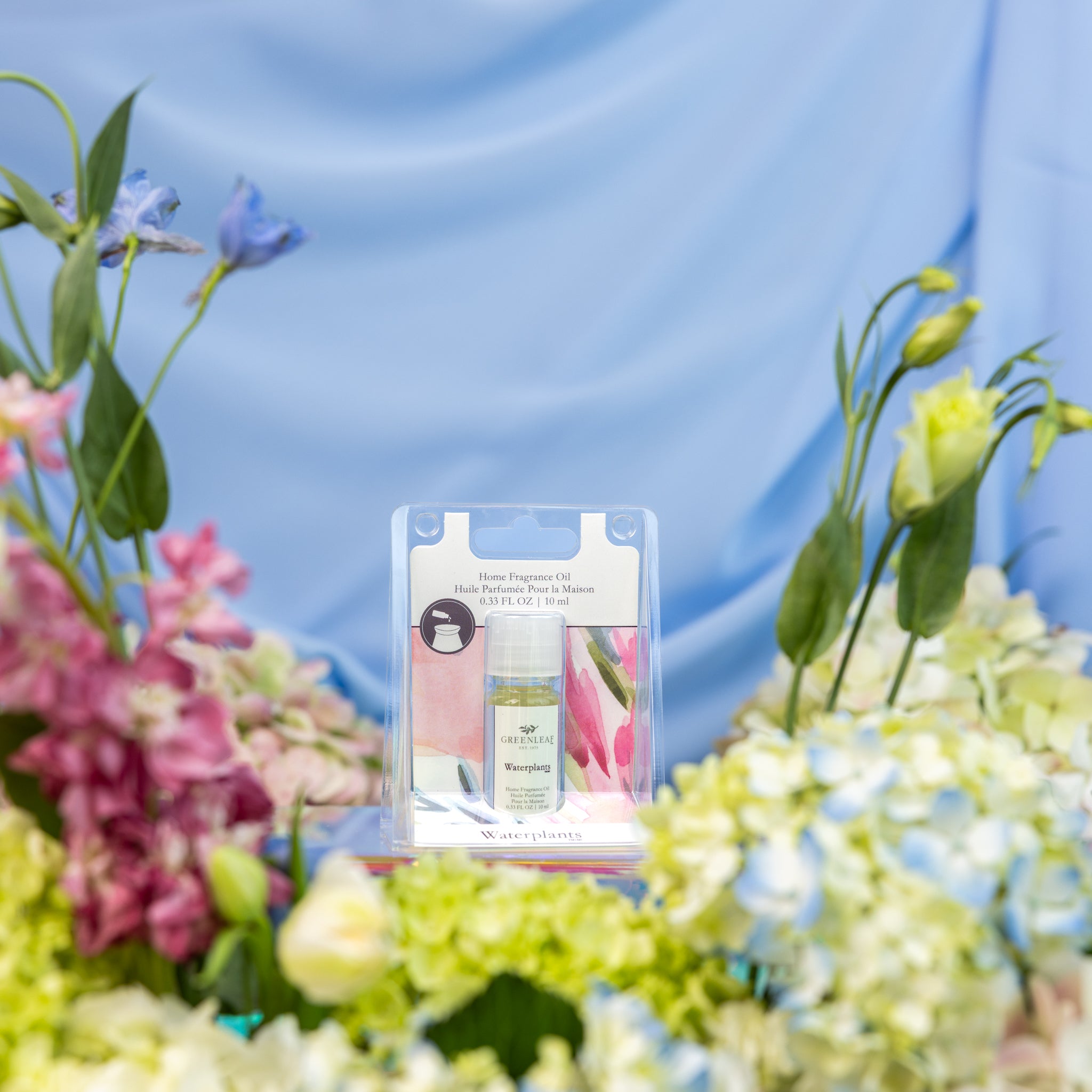 Greenleaf Gifts' Waterplants Home Fragrance Oil is displayed among vibrant flowers and waterplants, with a soft blue fabric backdrop, adding a touch of flameless fragrance elegance.
