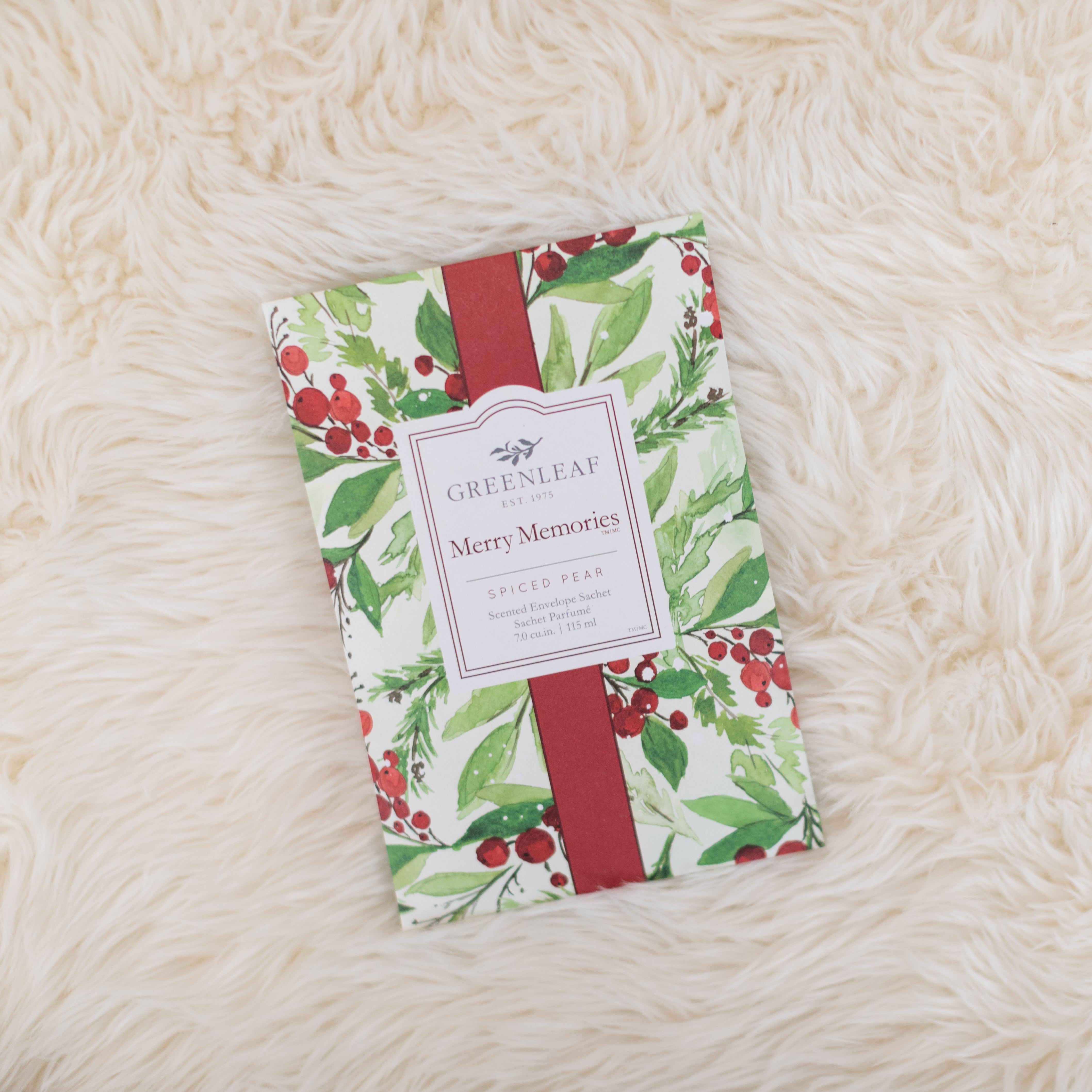 A Merry Memories Sachet from Greenleaf Gifts rests on a soft, cream-colored surface. Its packaging features holly and berry illustrations with a red ribbon design and offers a festive spiced pear scent.