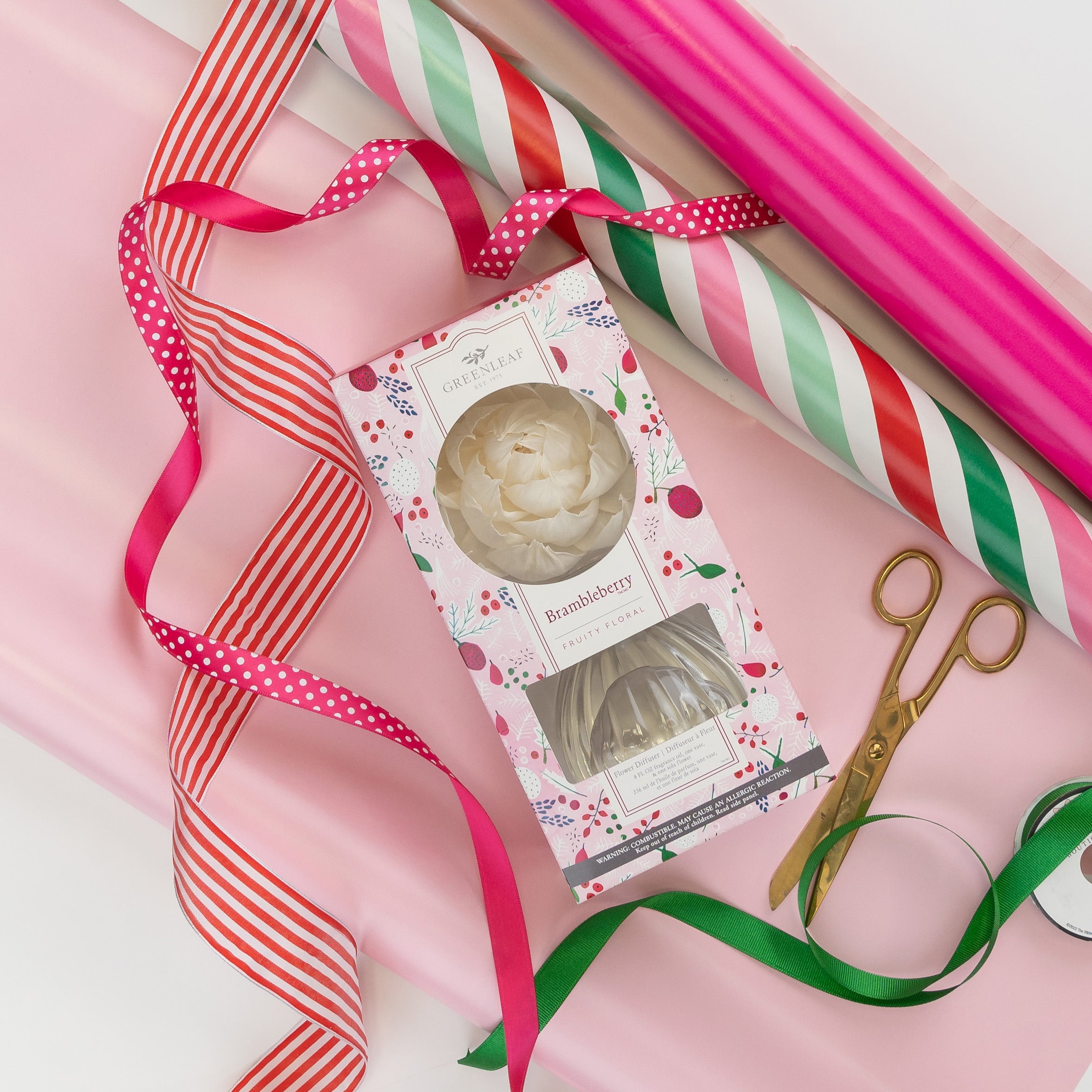 Gift wrapping supplies with pink and striped paper, red and green ribbons, gold scissors, and a Greenleaf Gifts Brambleberry Flower Diffuser arranged on a white surface.