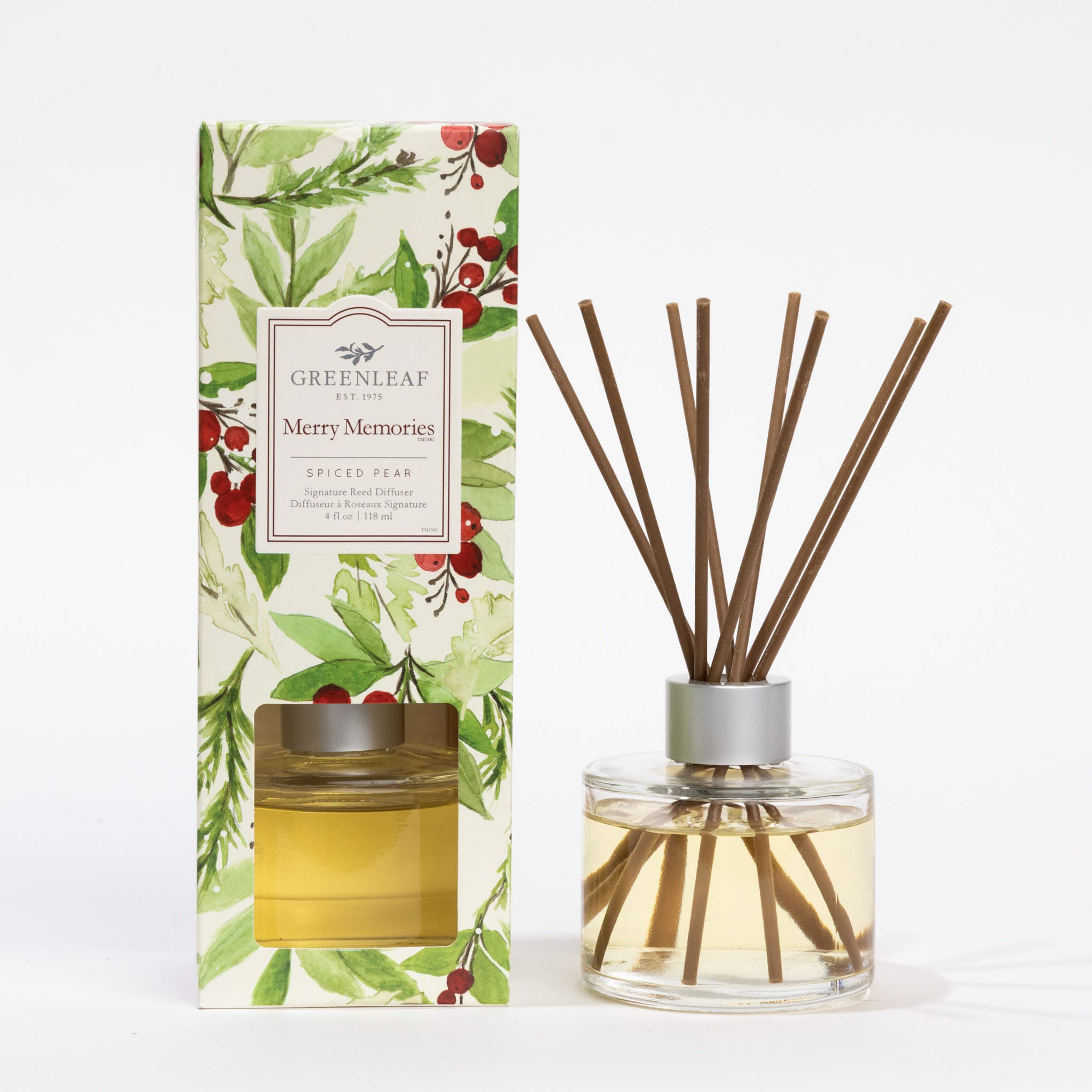 A glass Merry Memories Reed Diffuser filled with yellow liquid sits beside its box, adorned with green leaves and red berries. The label reads Greenleaf Gifts, offering a festive spiced pear fragrance.