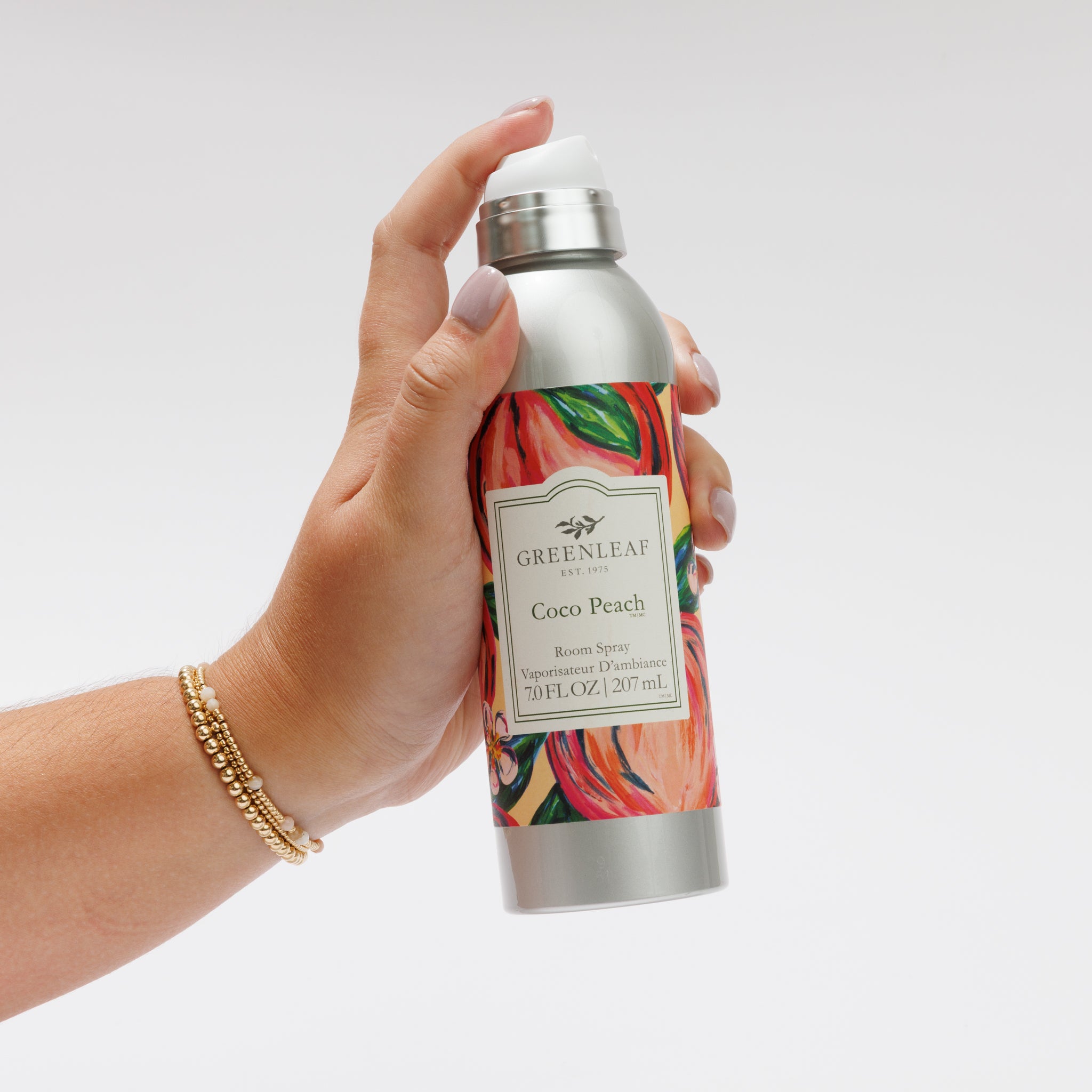 A hand holds a silver Greenleaf Gifts Coco Peach Room Spray with a colorful peach and green label, set against a plain white background.