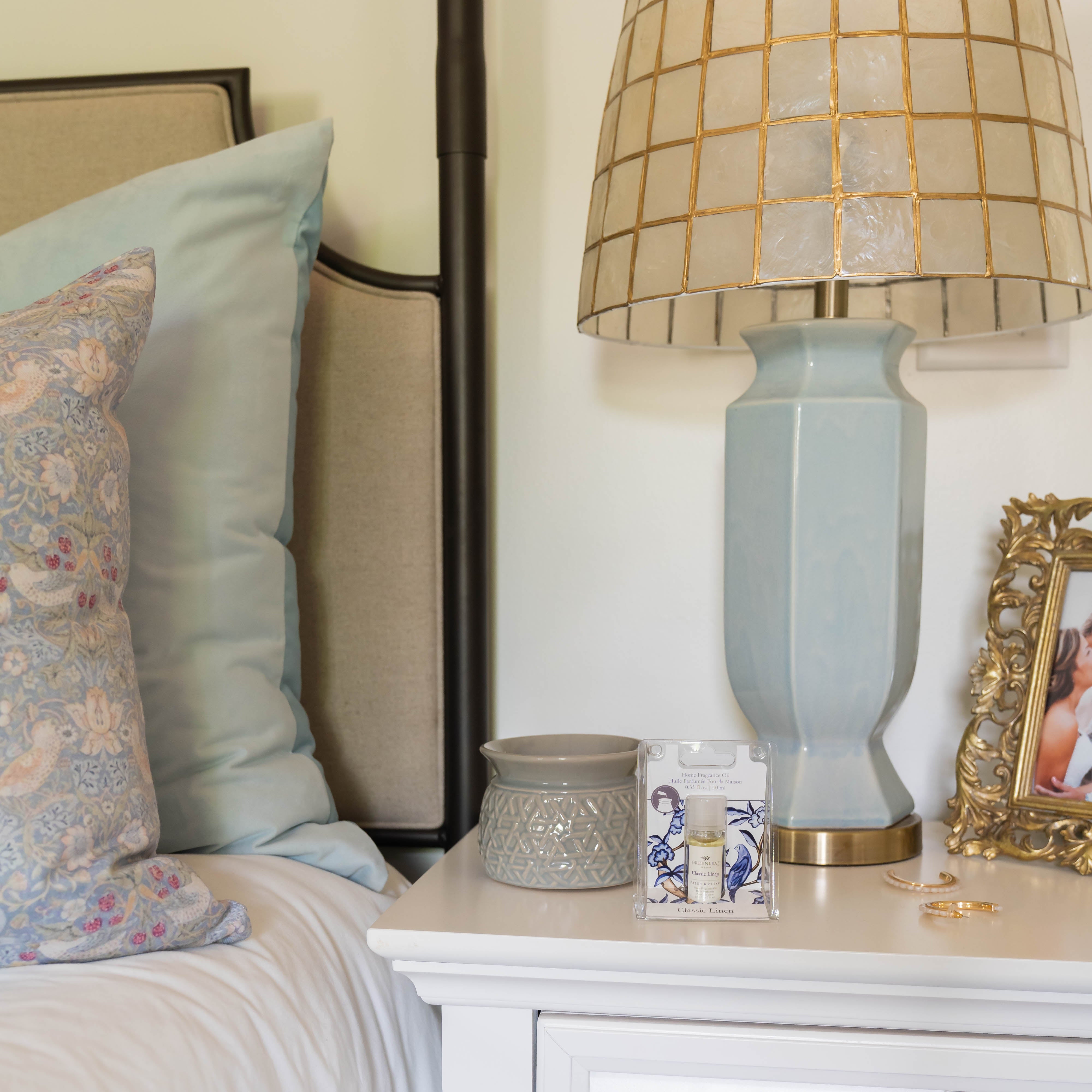 A bedside table holds a pale blue lamp, a small pot, a framed photo, jewelry, and beauty products. Next to it, the bed with light blue pillows and a floral cushion is scented by Greenleaf Gifts Classic Linen Home Fragrance Oil.