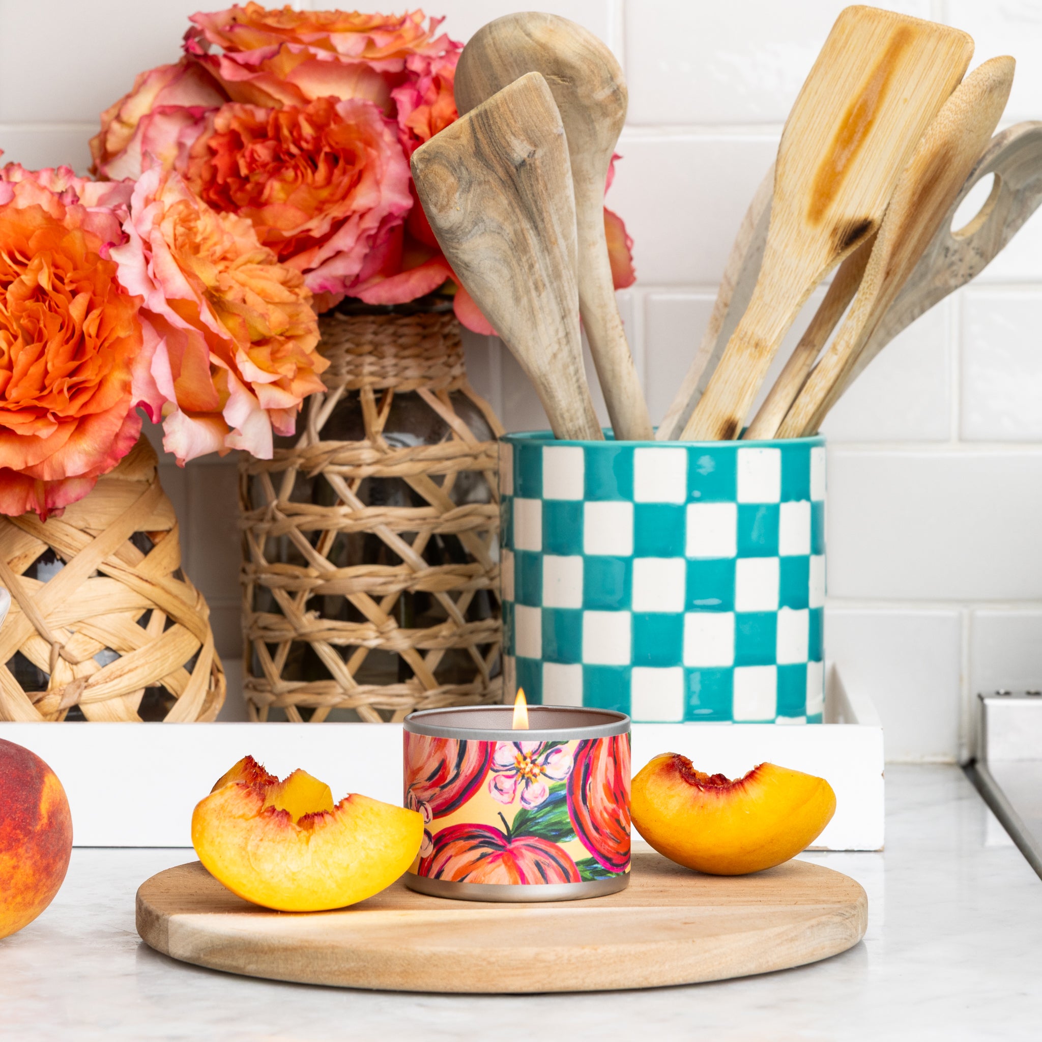 A Coco Peach Tin Candle by Greenleaf Gifts, sliced peaches, and a cutting board rest on a white counter. Nearby, orange flowers in woven vases and wooden utensils in a turquoise checkered holder bring Cocoa Peach charm.