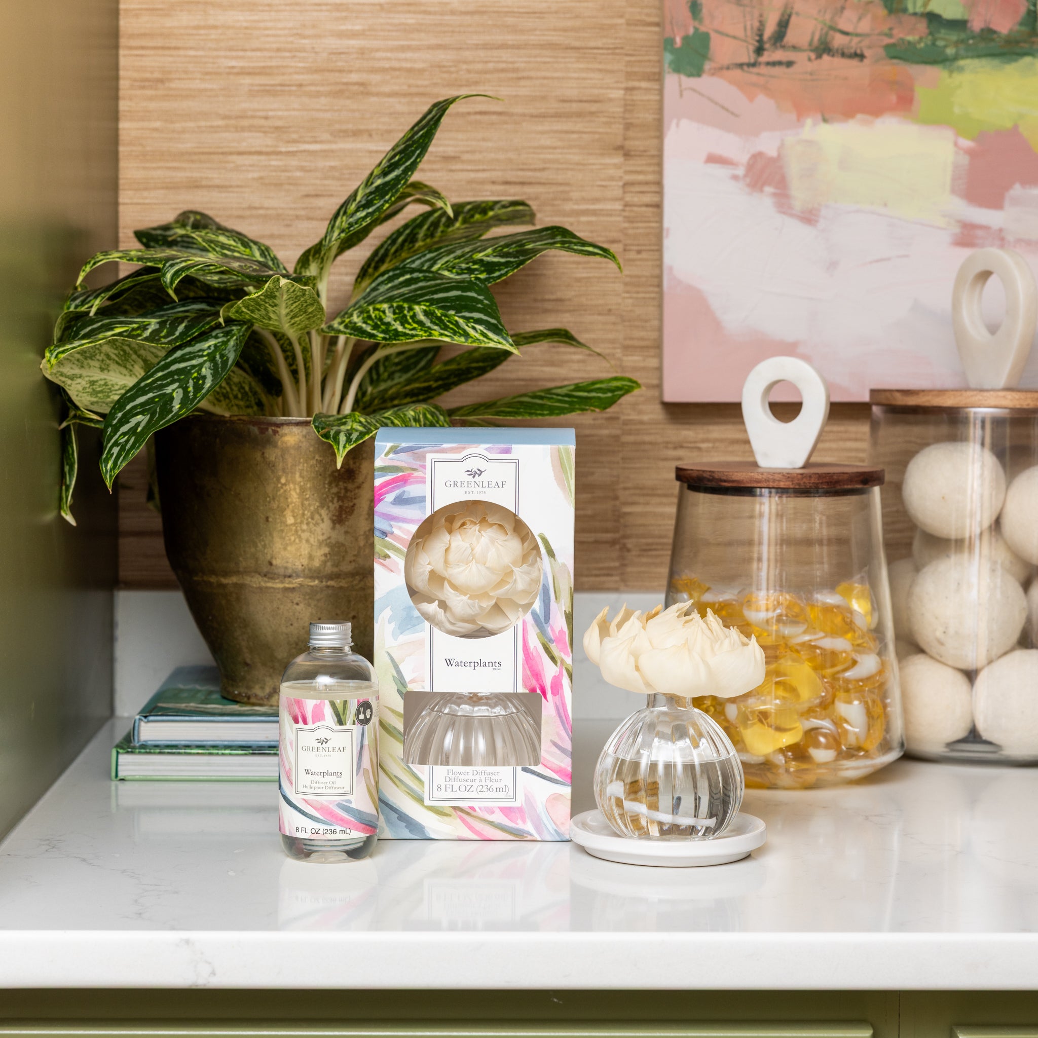 A countertop displays a potted plant, a boxed Waterplants Diffuser Oil by Greenleaf Gifts, a glass diffuser with a cream-colored flower, an open Greenleaf Gifts Diffuser Oil bottle, two jars filled with yellow and white items, and art in the background.