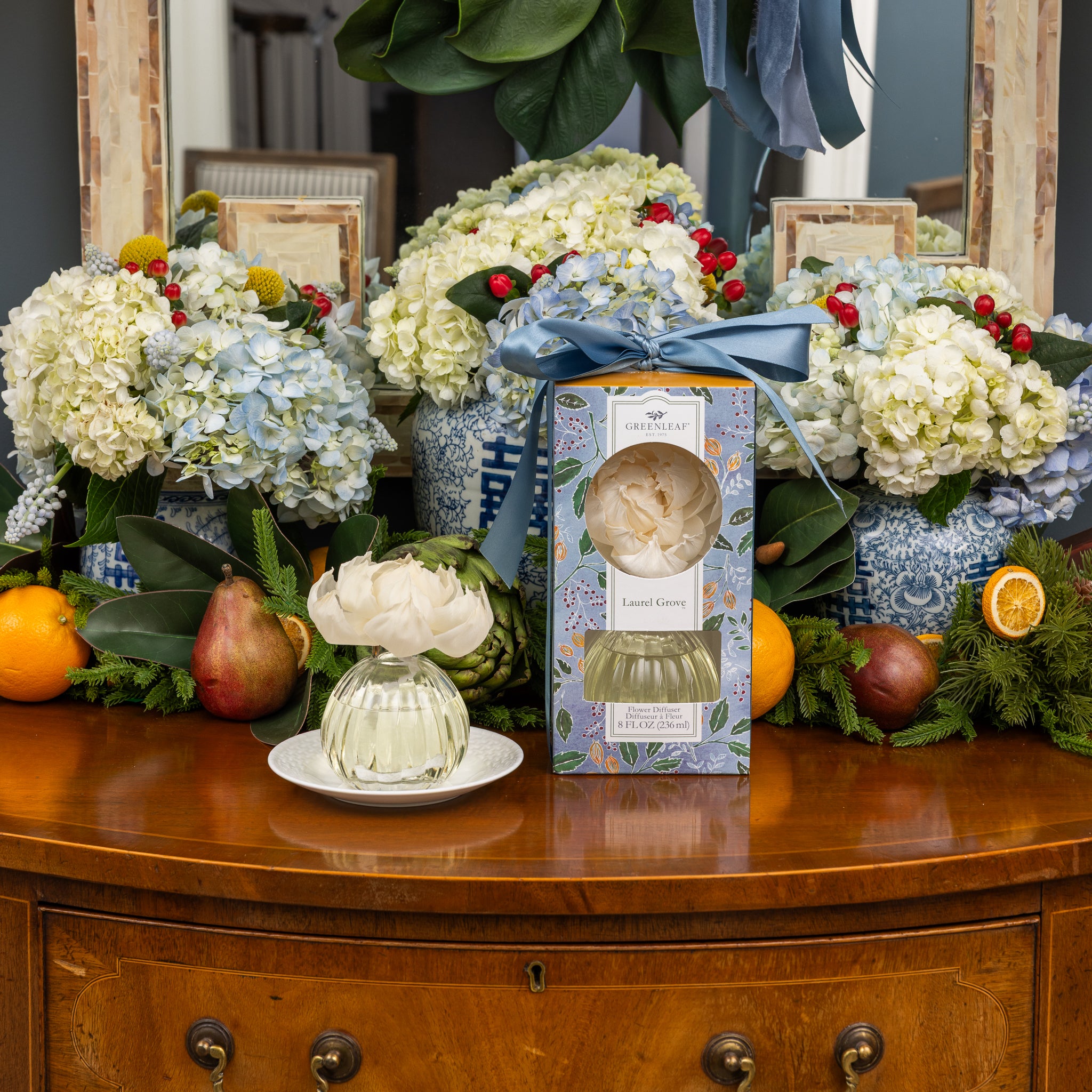 A decorative wooden dresser displays assorted fruits, a Greenleaf Gifts Laurel Grove Flower Diffuser set, and a floral arrangement with blue hydrangeas and a sola paper flower, all reflected in the mirror behind.