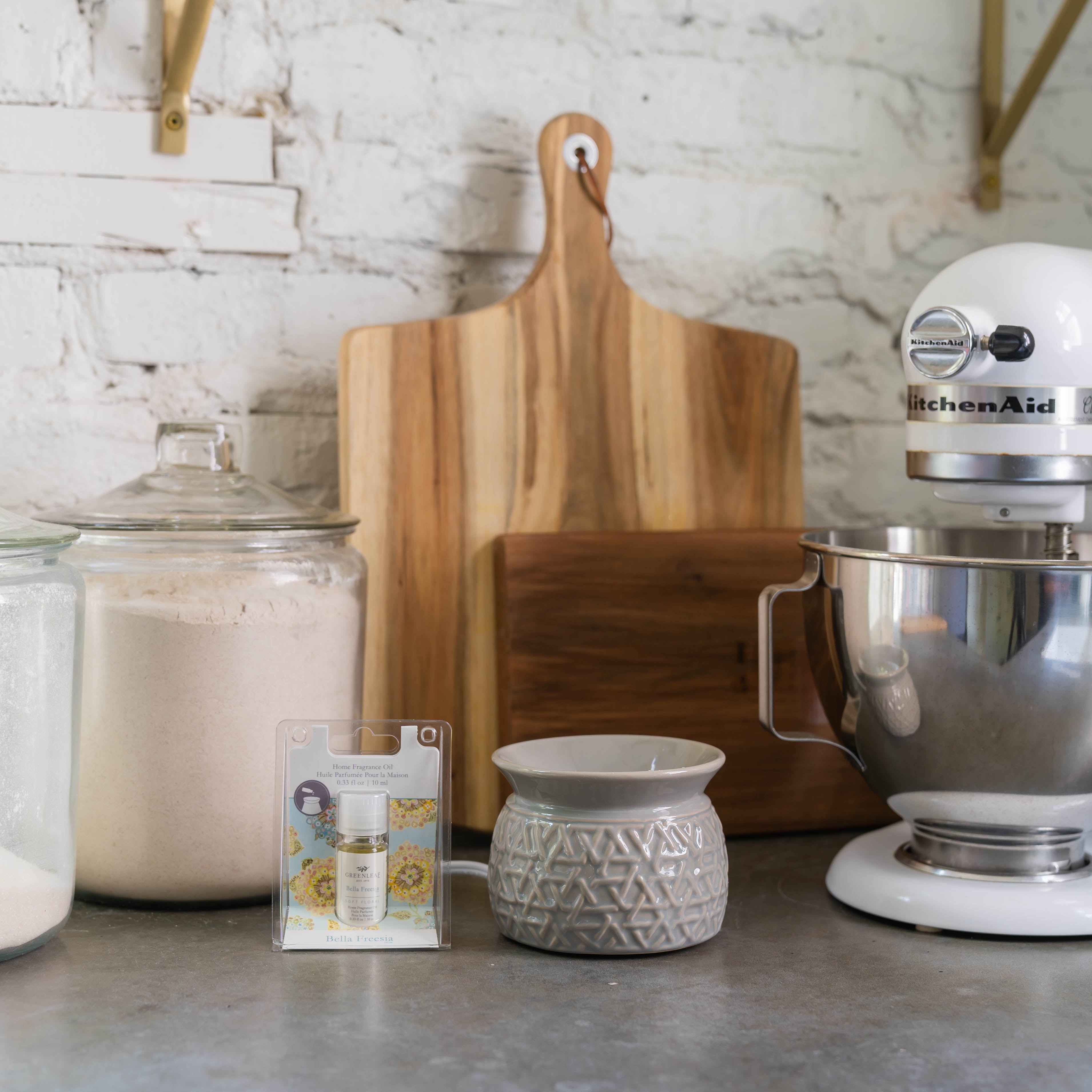 A kitchen counter with a stand mixer, bowls, jars, and cake decoration gel in front of a white brick wall is subtly enhanced by the scent of Greenleaf Gifts’ Bella Freesia Home Fragrance Oil.