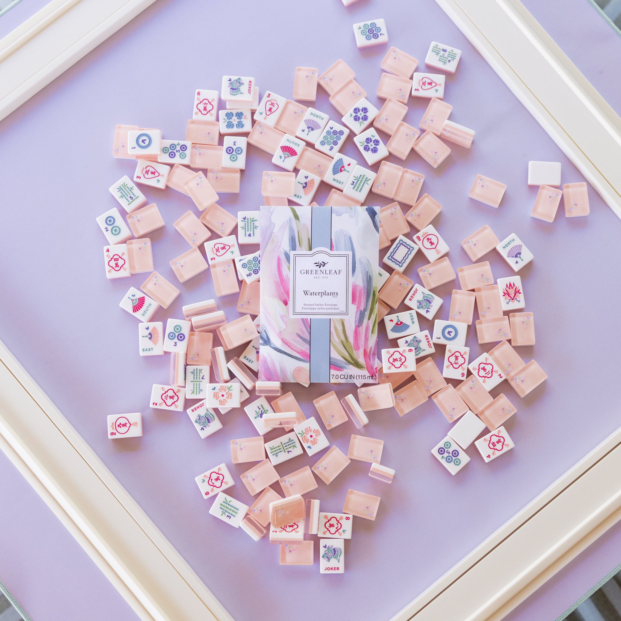 A Greenleaf Gifts Waterplants Sachet sits amid pastel pink mahjong tiles with colorful symbols, arranged on a lavender surface in a white frame—a charming display for this long-lasting air freshener.