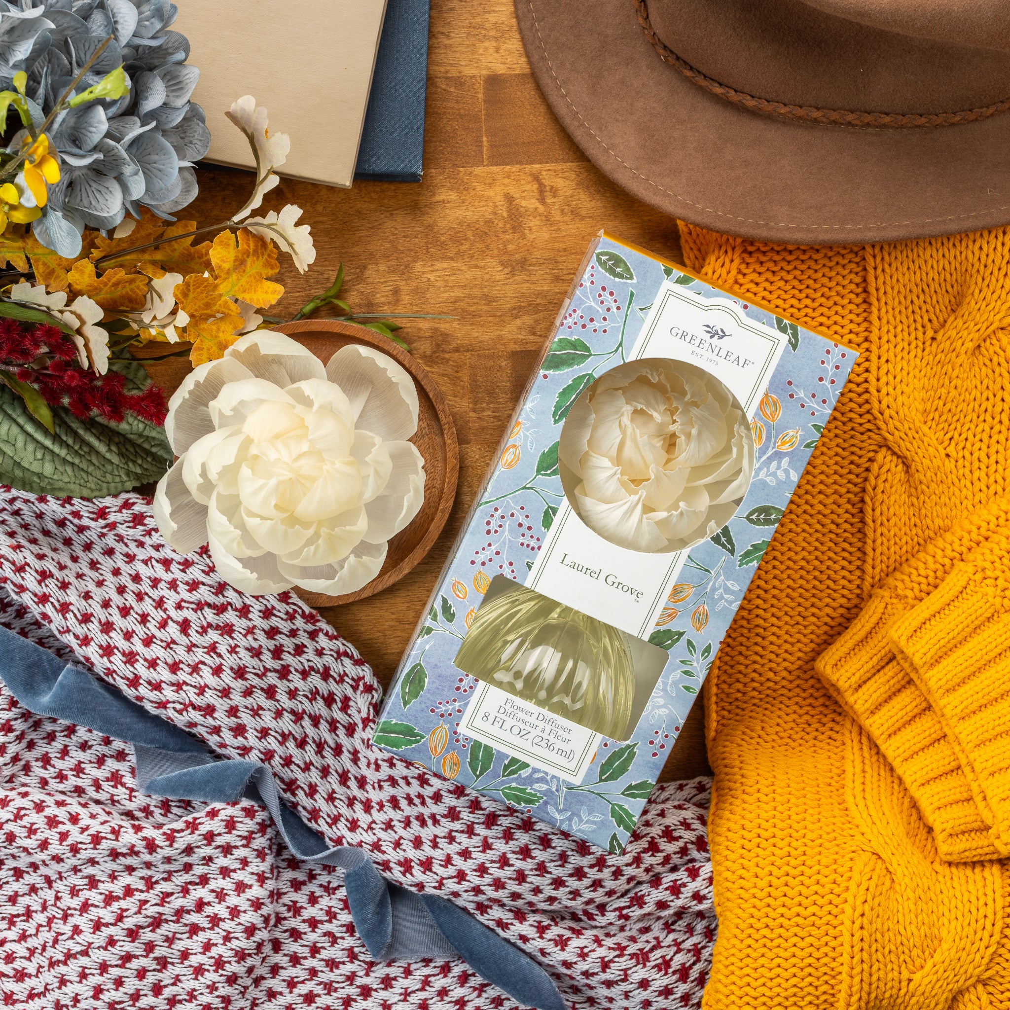 A Greenleaf Gifts Laurel Grove Flower Diffuser in floral packaging sits on a wooden table with a yellow knit sweater, red-and-white scarf, faux flowers, and a brown hat, filling the room with fresh Greenleaf fragrance.