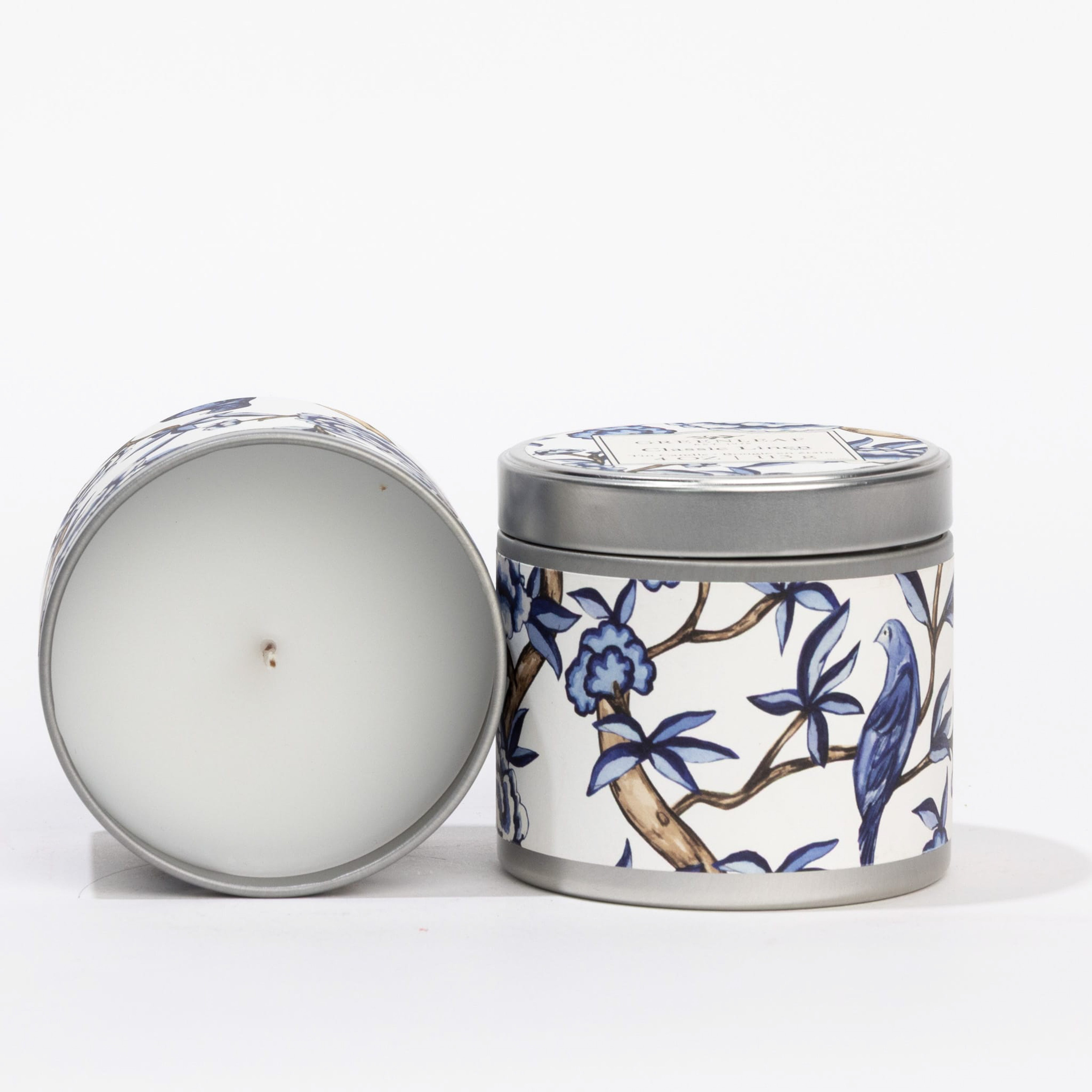 Greenleaf Gifts’ Classic Linen Tin Candle features a blue floral and bird design with silver lid; one stands upright, the other on its side, displaying white wax and centered wick—perfect for adding charm to any space.