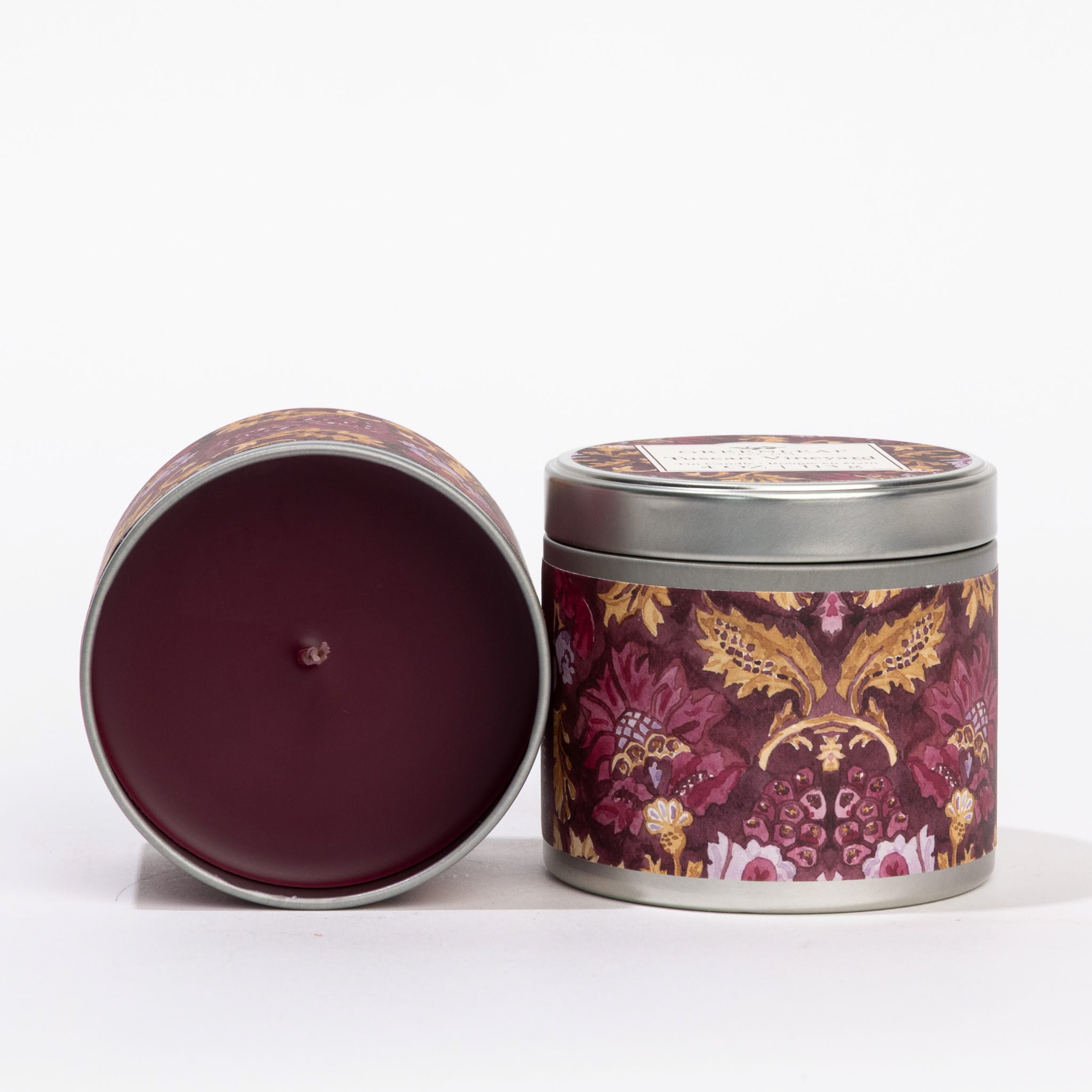 Two Greenleaf Gifts Tuscan Vineyard Tin Candles with maroon, gold, and pink designs—one upright with a lid, one on its side displaying the candle inside—bring the charm of a Tuscan vineyard wherever you are.