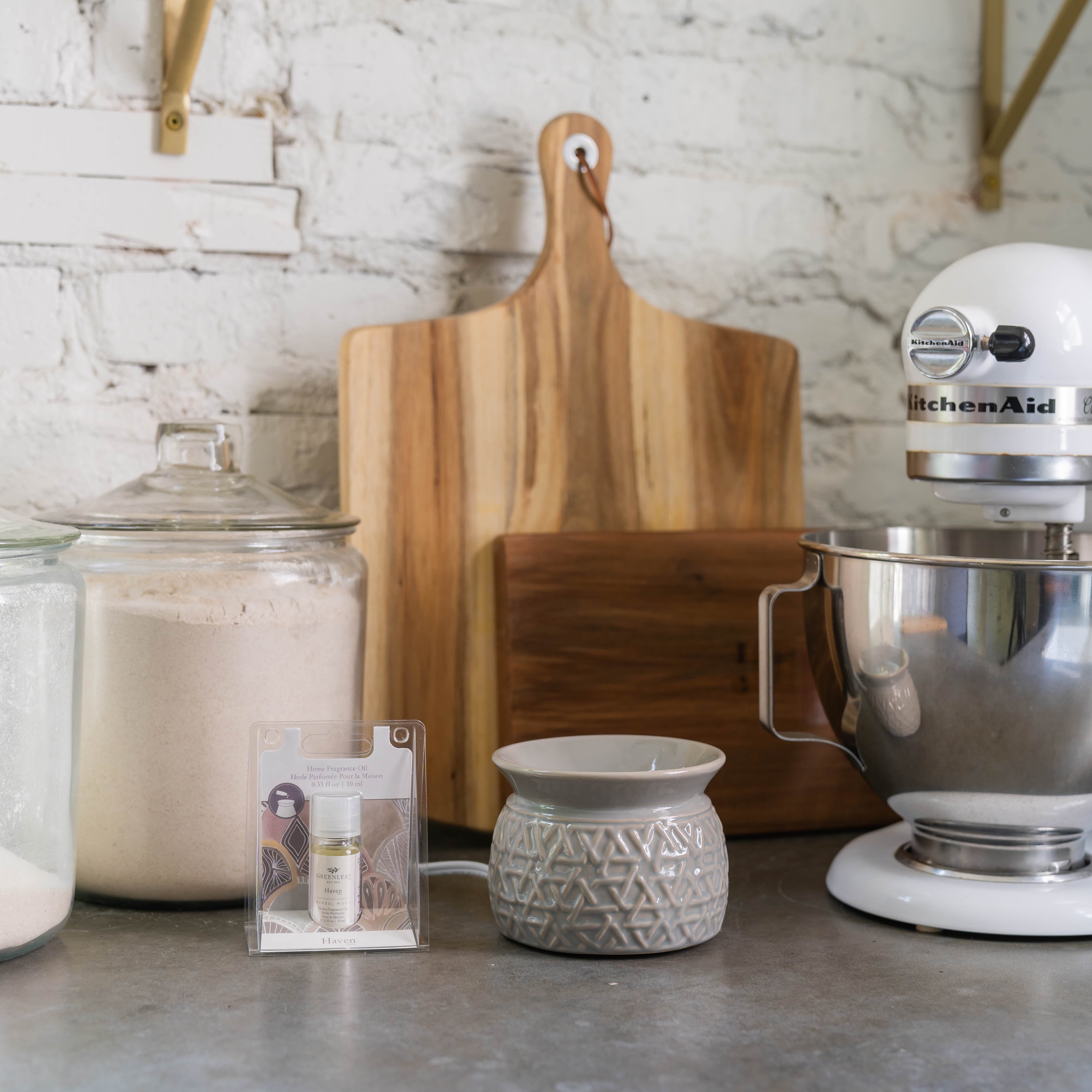 A kitchen counter with flour and sugar jars, a wooden cutting board, a ceramic flameless fragrance warmer, Greenleaf Gifts Haven Home Fragrance Oil, and a white KitchenAid stand mixer against a white brick wall.