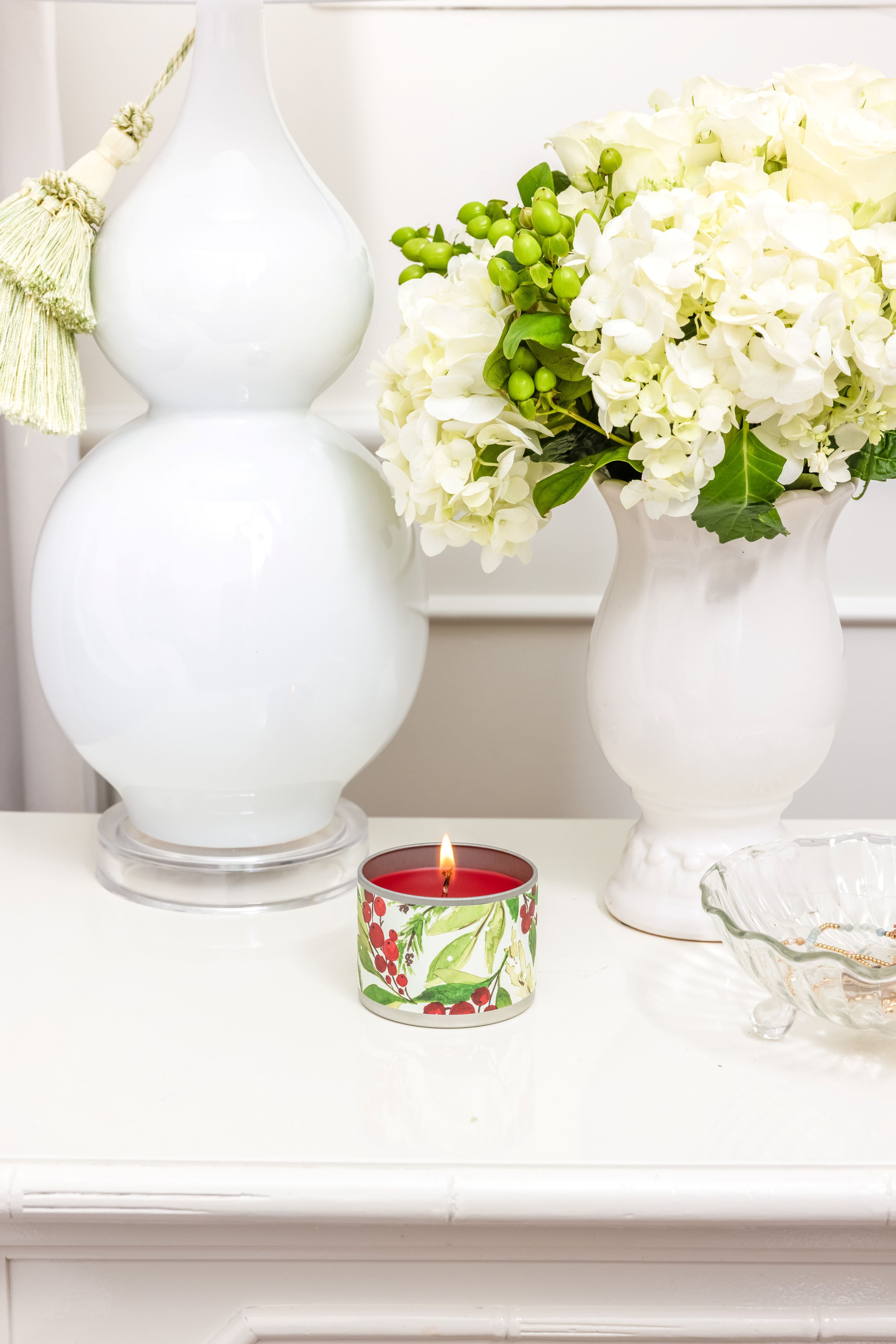 The Merry Memories Tin Candle by Greenleaf Gifts, in a floral-patterned tin, sits on a white table beside a white lamp, a vase of white flowers with greenery, and a clear decorative dish.