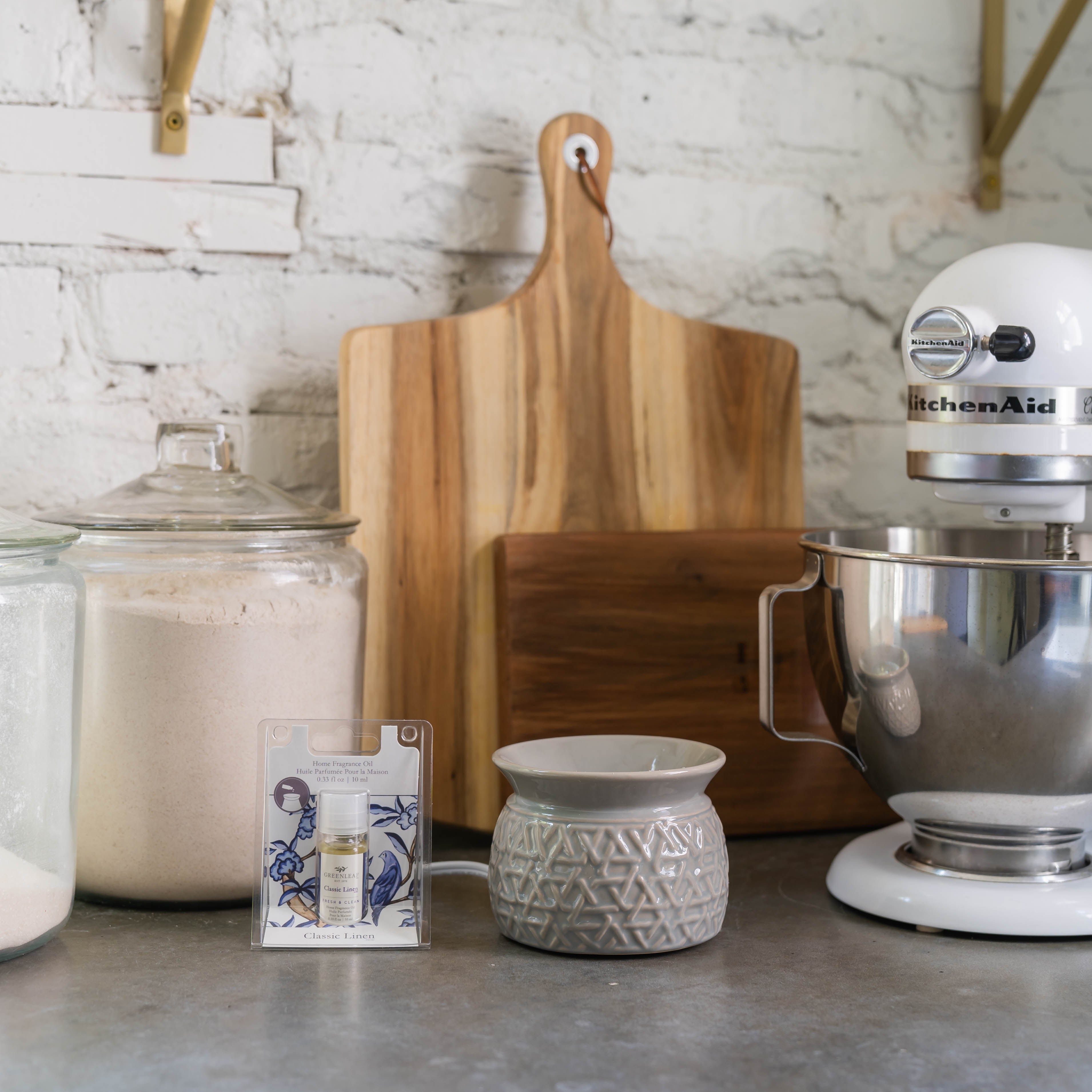 A kitchen counter features a KitchenAid mixer, wooden cutting board, glass jars of flour and sugar, a flameless wax warmer with Greenleaf Gifts Classic Linen Home Fragrance Oil, and a decorative card—all set against a white brick wall.