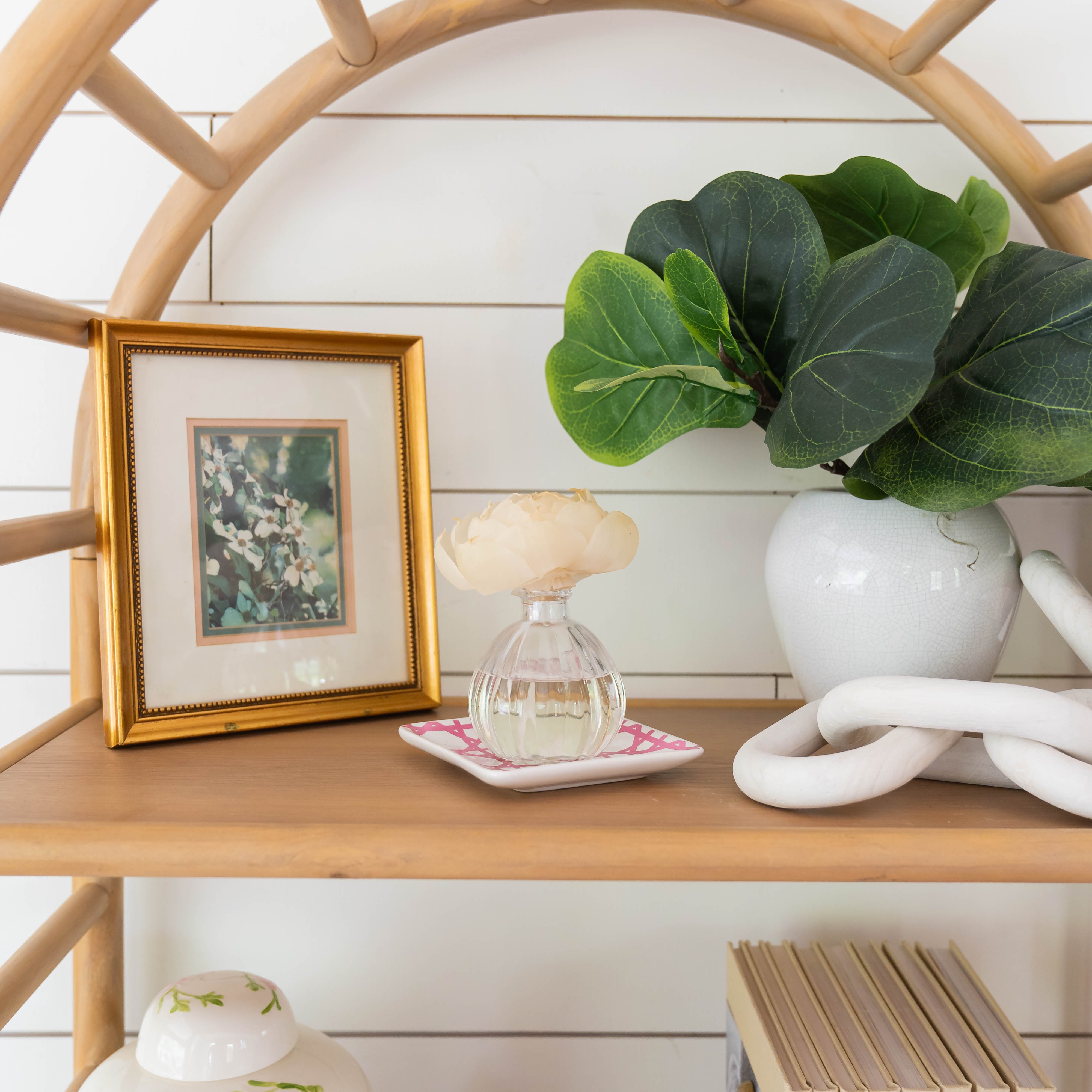 A wooden shelf holds a framed picture, a vase with green leaves, the Greenleaf Gifts Flower Diffuser-Classic Linen with a light flower, a decorative white knot, and books. The background features white shiplap paneling.