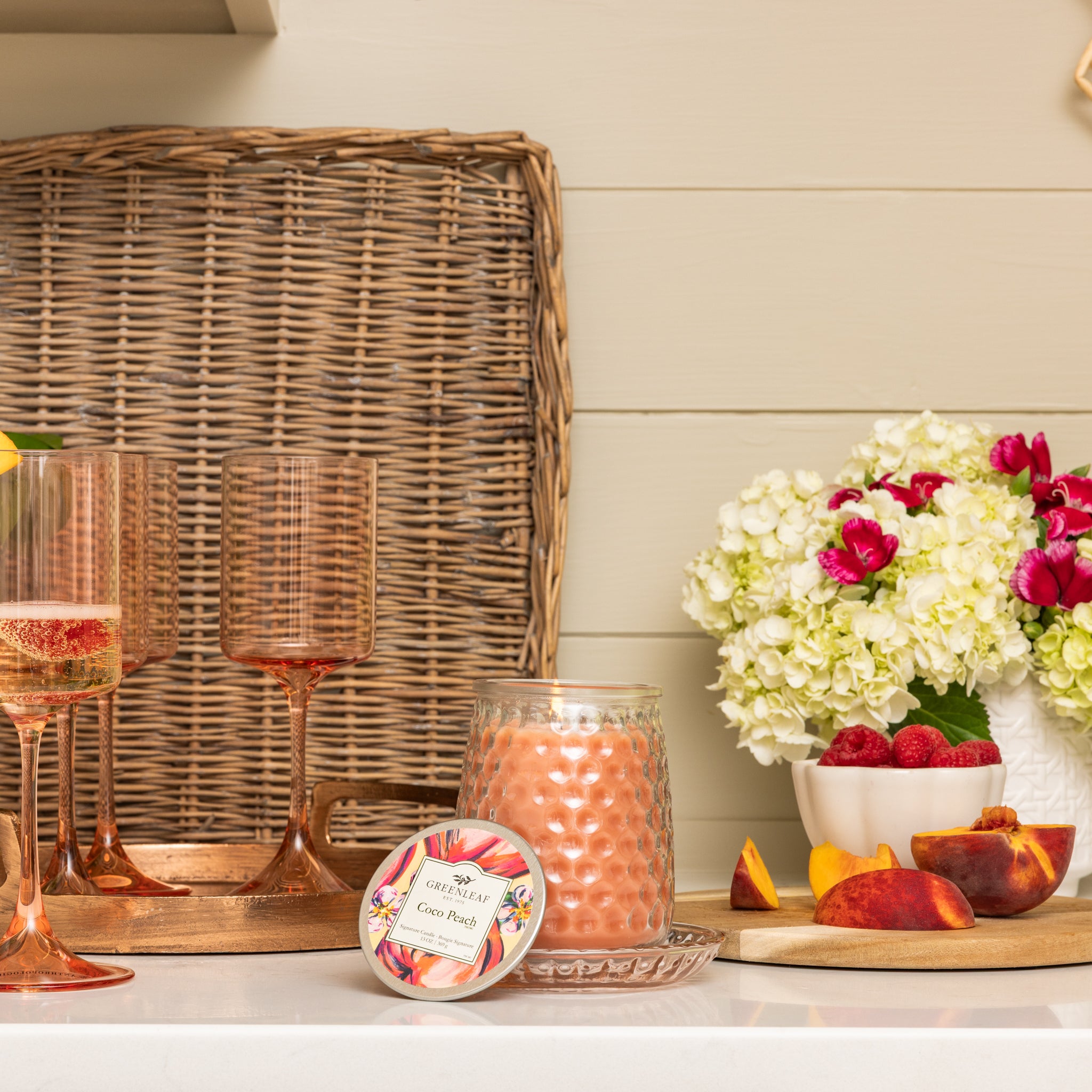 A kitchen counter displays peach glasses, a lit Greenleaf Gifts Coco Peach Signature Candle, a floral-labeled tin, a tray, white hydrangeas with red flowers, and a bowl of strawberries and peach slices—infused with Greenleaf fragrances.