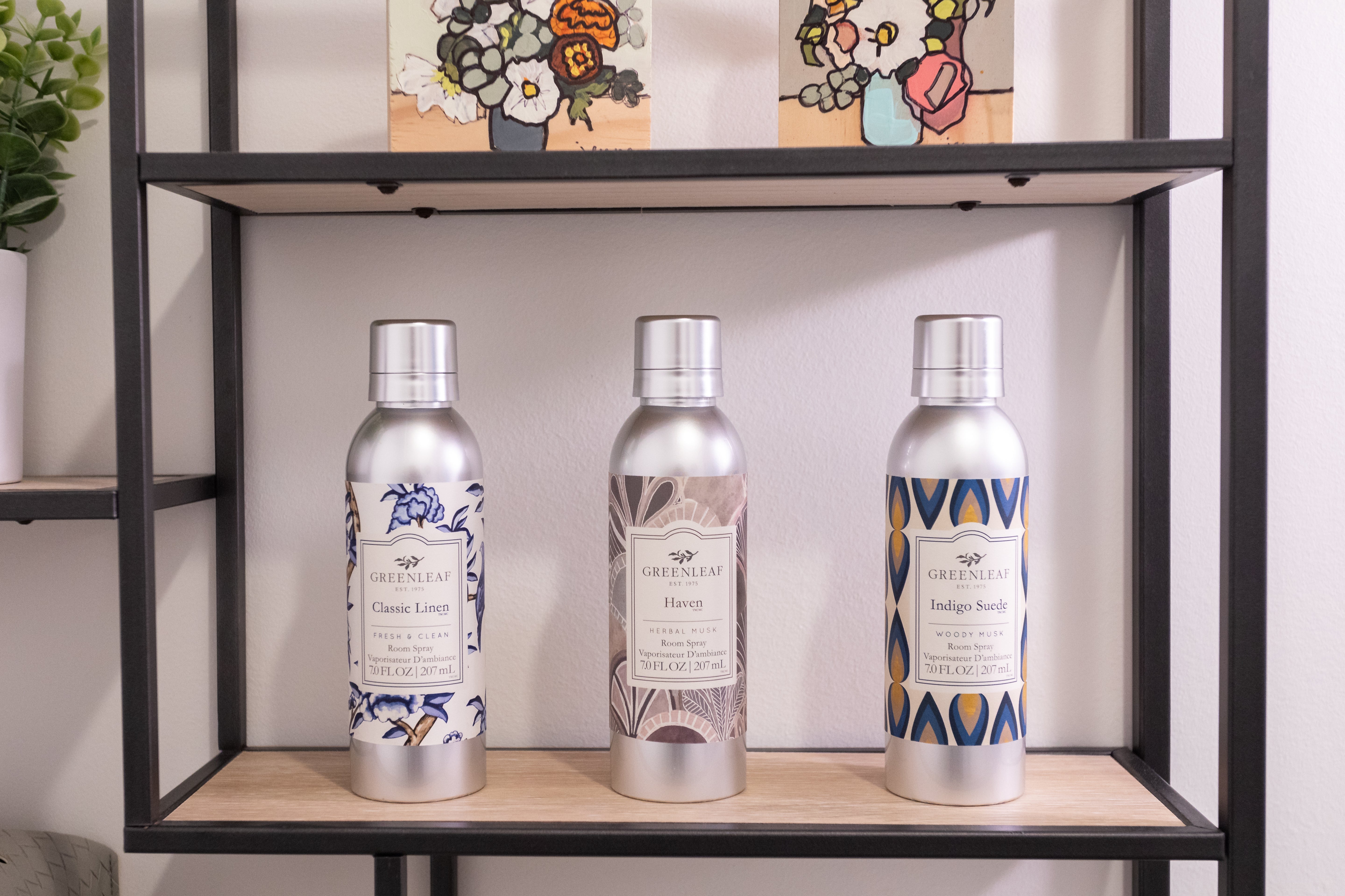 Three Greenleaf Gifts Classic Linen Room Sprays are displayed on a shelf, each with a decorative label. Above the sprays are small colorful paintings and a potted plant.