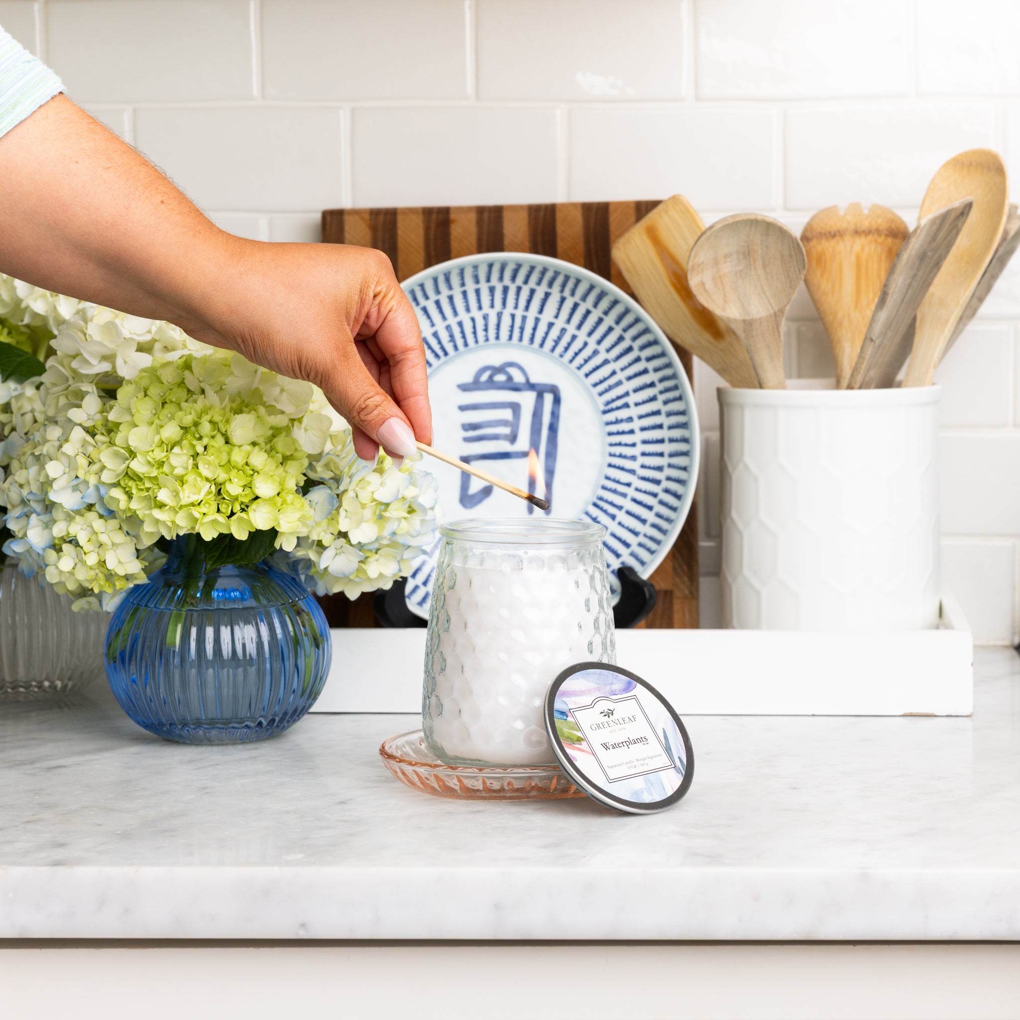 A person’s hand lights a Greenleaf Gifts Waterplants Signature Candle in a glass jar on a kitchen counter, beside a vase of flowers, other Greenleaf fragrances, a plate, and wooden utensils; the candle lid sits nearby on a small glass plate.